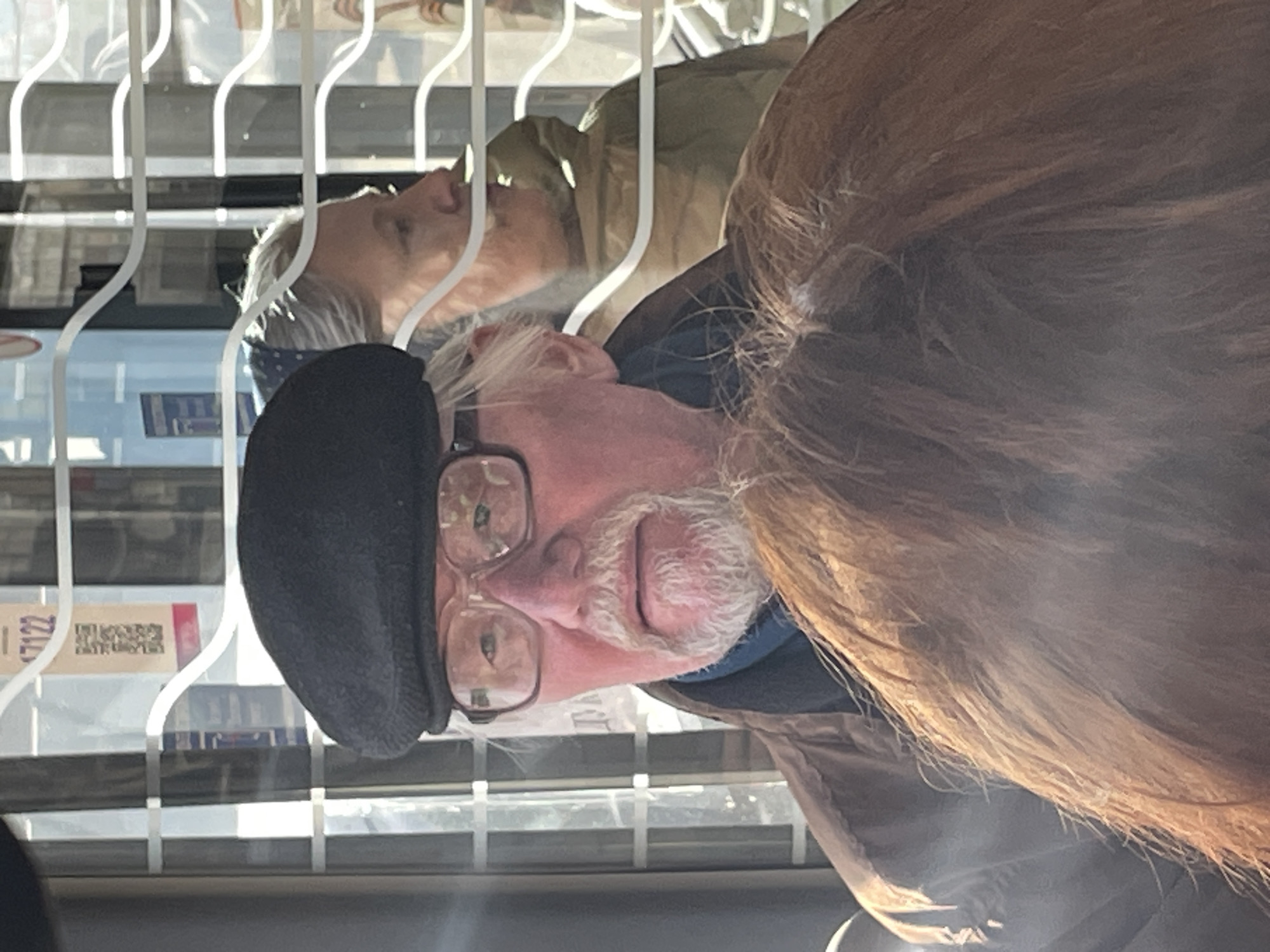 An older man wearing glasses and a black beret is looking at objects behind a wire display rack. A second person can be seen, in a different plane, reflected in the glass of the display rack. A sticker with numbers, "17122" is partially visible on the rack. The image orientation is turned 90 degrees counterclockwise. The location is inferred to be Krasnoyarsk, Russia based on the tags but is not visible in the image.