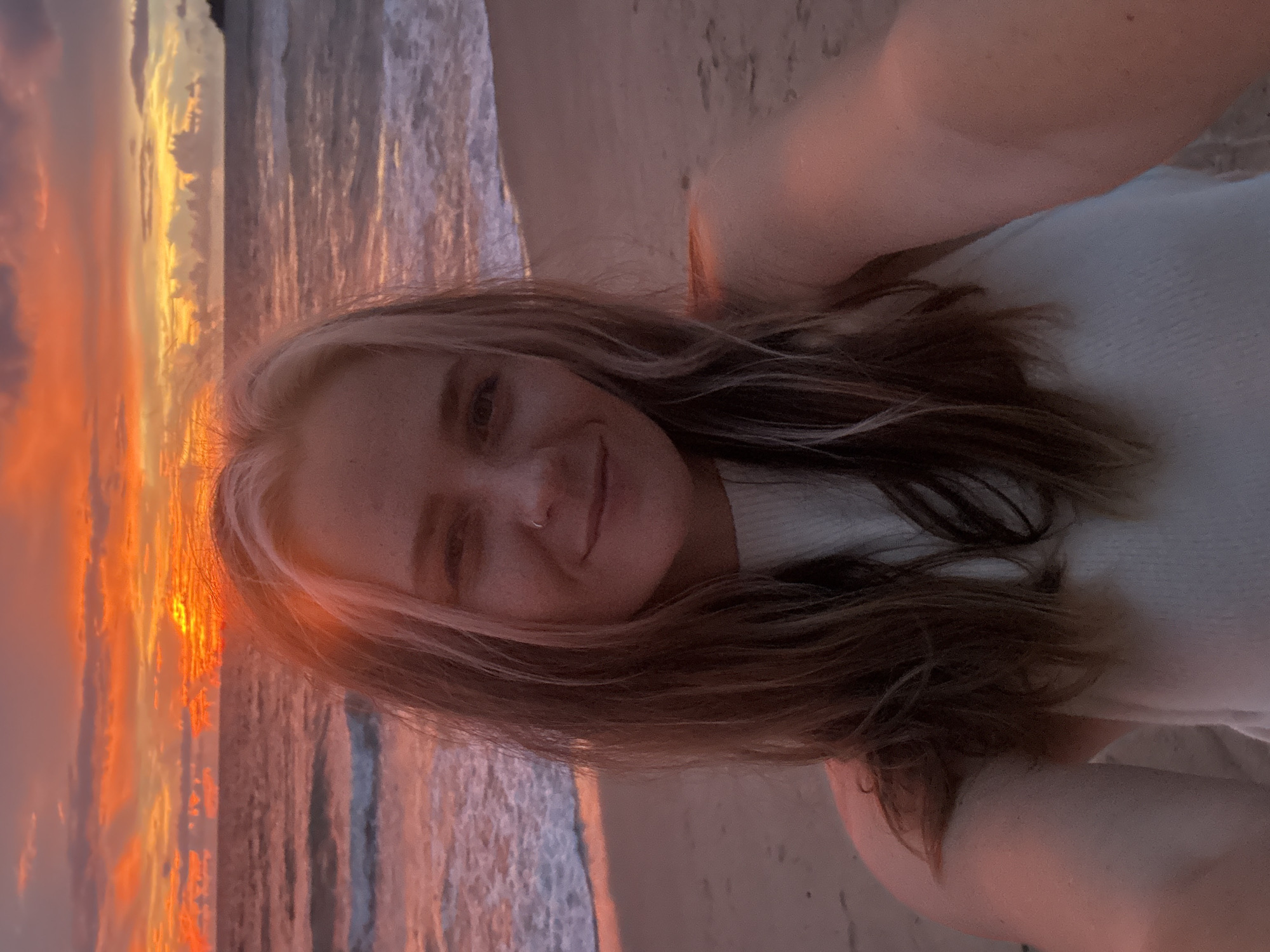A young woman with blonde hair, wearing a white top, takes a selfie at the beach during sunset. The sky is a vibrant mix of orange, yellow, and pink hues, reflecting on the ocean. The waves are gently rolling onto the shore, creating a peaceful atmosphere. The woman is smiling and winking at the camera, her face illuminated by the warm glow of the setting sun. Her hair is long and wavy, framing her face. A small nose ring is visible on her right nostril. The overall mood is relaxed and joyful, capturing a beautiful moment at the end of the day.