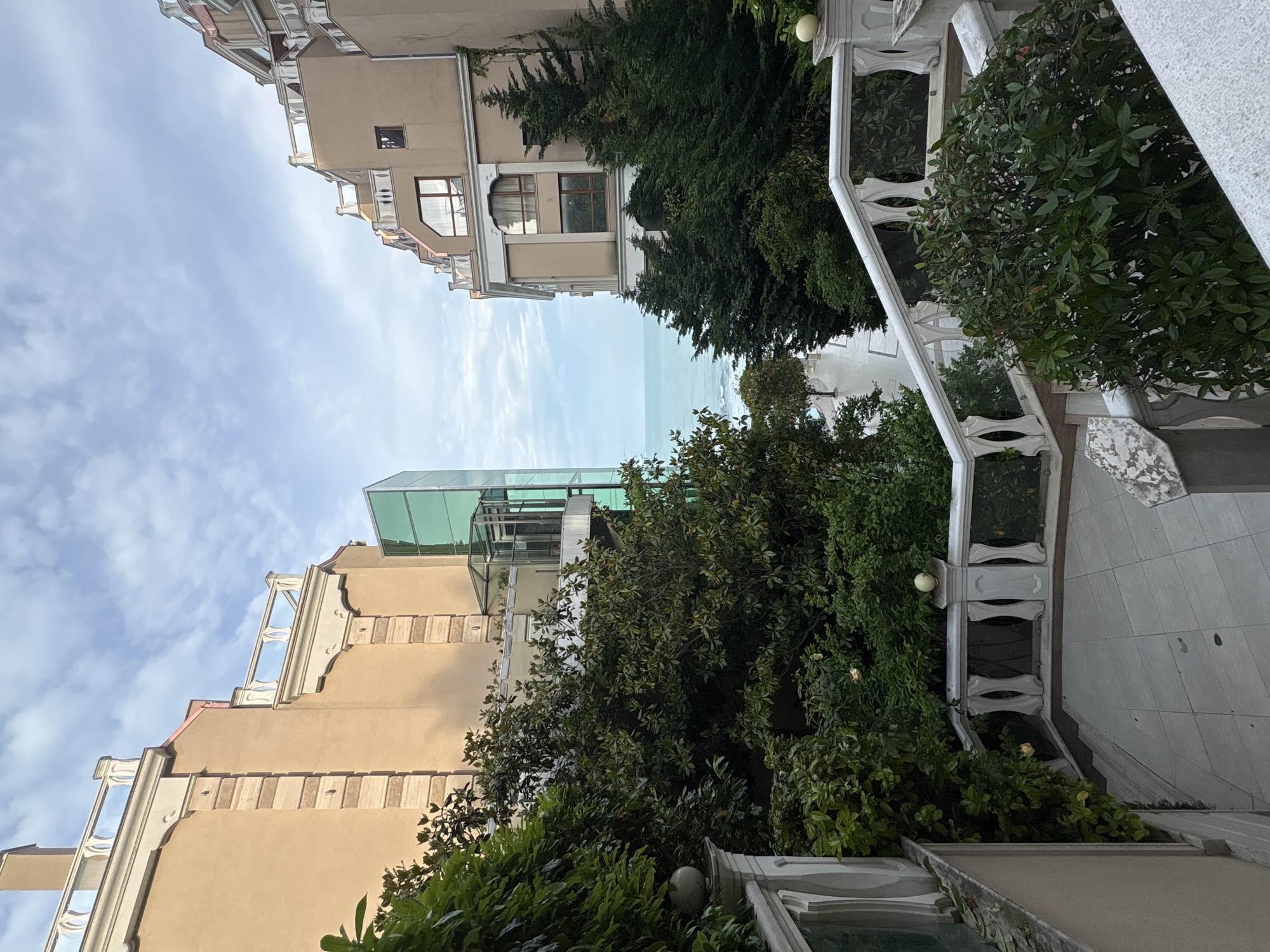 The image displays an outdoor scene looking down a narrow passage between two multi-story buildings towards a body of water. The two buildings, flanking the view, are constructed with beige or light brown facades. They feature numerous windows, balconies adorned with decorative white balustrades, and intricate architectural moldings.
Lush green trees and dense shrubs are abundant, filling the lower and mid-ground spaces between the buildings and lining a pathway on the right. On the right side, a prominent white stone balustrade with ornate oval cutouts angles downwards, suggesting a staircase or terraced walkway. A spherical light fixture is mounted on one of the balustrade posts. On the upper left, a modern glass and metal structure, possibly an elevator shaft or a rooftop enclosure, rises between the buildings.
In the distance, beyond the foliage and structures, a calm, light blue expanse of water is visible, indicating a coastal or lakeside setting. The sky above is partly cloudy with visible patches of blue.
The time of day appears to be daytime, with soft, natural lighting suggesting either morning or late afternoon. No people or specific activities are depicted, contributing to a serene and tranquil atmosphere. There is no visible text in the image.