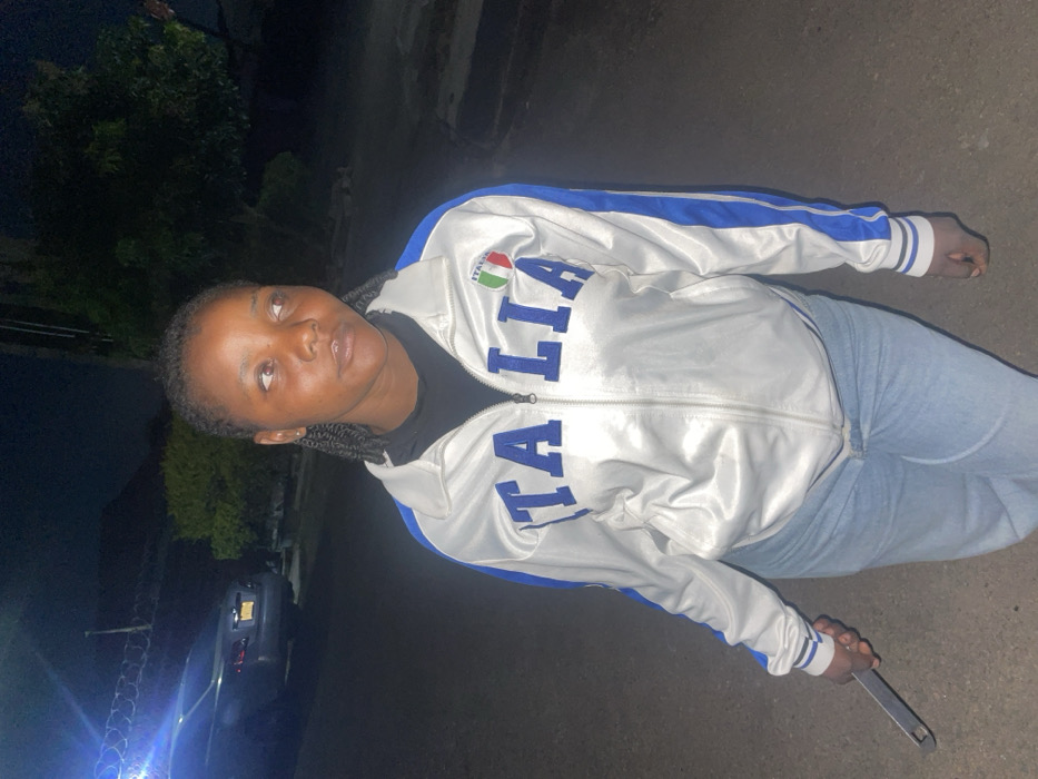 A woman in a white tracksuit with "ITALIA" printed on the front stands on a dark street. She's wearing light blue jeans and holding a long, silver wrench in her right hand. The surrounding area is dimly lit, with sparse vegetation and a car visible in the background. The overall mood is serious, given the woman's expression and the presence of the wrench.