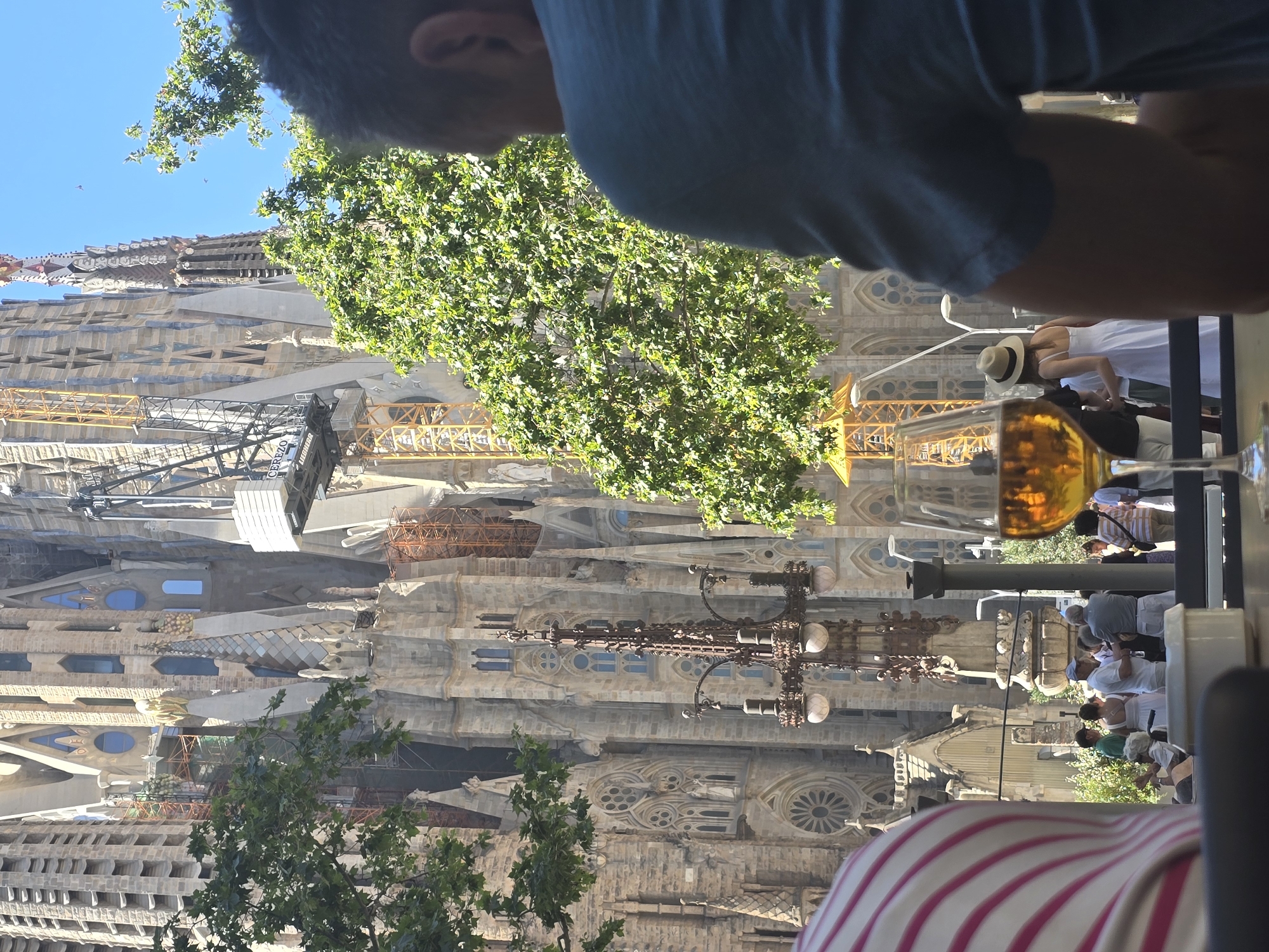 The image depicts a view of the Sagrada Familia in Barcelona, Spain. In the foreground, a glass of amber liquid, likely beer or wine, sits on a table. To the left, the back of a person wearing a striped shirt is visible. In the midground, a group of people are gathered outside the Sagrada Familia, some standing and talking, one person sitting in a wheelchair. A lamppost with multiple globes stands prominently. Behind them, the iconic facade of the Sagrada Familia is visible, with construction cranes and scaffolding indicating ongoing work. Lush green trees frame the scene, with bright sunlight filtering through their leaves. The sky is clear and blue. The overall impression is a relaxed, sunny day at a major tourist attraction.