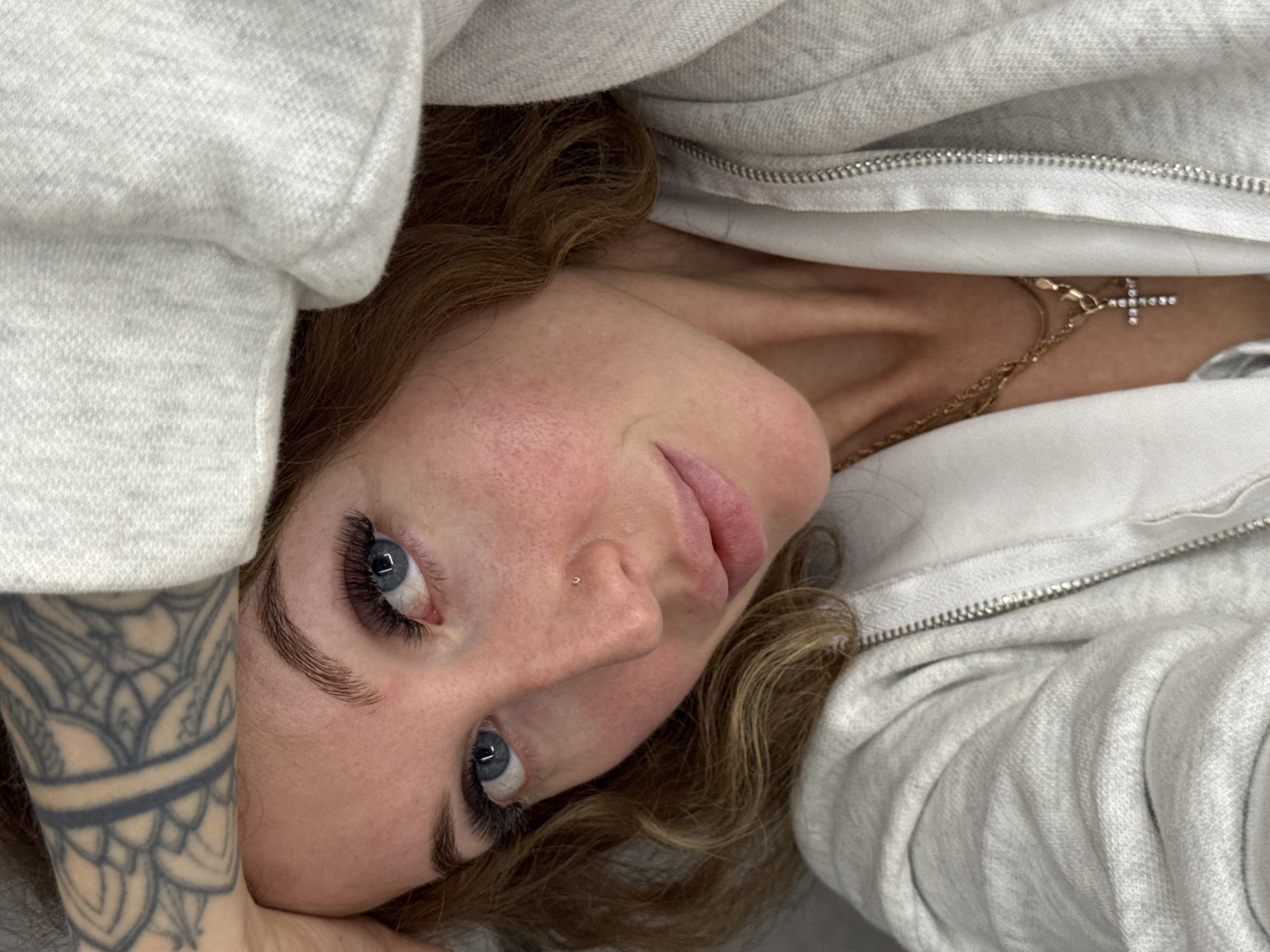The image captures a close-up, slightly rotated overhead view of a person, likely a woman, with fair skin, light blue eyes, and reddish-brown, wavy hair. She is lying down, looking towards the camera with a calm, neutral expression. Her eyes are open, revealing prominent, dark eyelashes, possibly extensions, and neatly shaped eyebrows. A small, sparkly stud piercing is visible on her nose. Her lips are slightly parted.
She is wearing a light grey hooded sweatshirt or jacket, the fabric of which frames much of her face and body. A silver zipper is visible along the opening of the garment near her neck. Around her neck, a thin gold chain necklace with a small, sparkling cross pendant is visible. On her left forearm, a black patterned tattoo featuring intricate designs is prominently displayed. The overall setting appears intimate and indoor, with soft, even lighting. There are no other discernible objects or specific location cues within the frame, aside from the person and her immediate surroundings of clothing. No text is visible. This scene is contextualized within Chaikovsky, Russia.