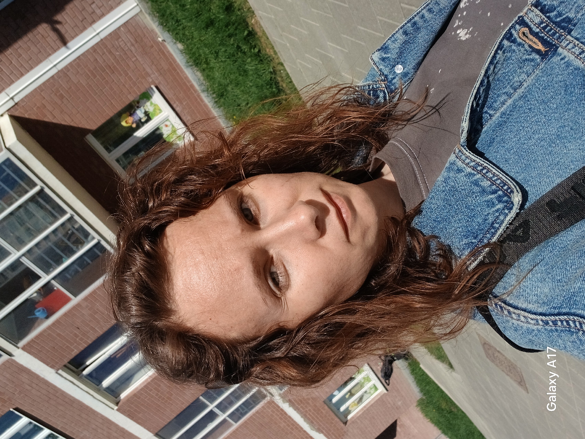A person is in the frame, wearing a dark-colored t-shirt and a blue denim jacket. The person's hair is reddish-brown and wavy. A black strap is visible across their torso. The background consists of a brick building with large windows, a patch of green grass, and a paved walkway. The picture was taken with a Galaxy A17, according to text in the lower right corner. This photograph appears to be taken in Yekaterinburg, Russia.