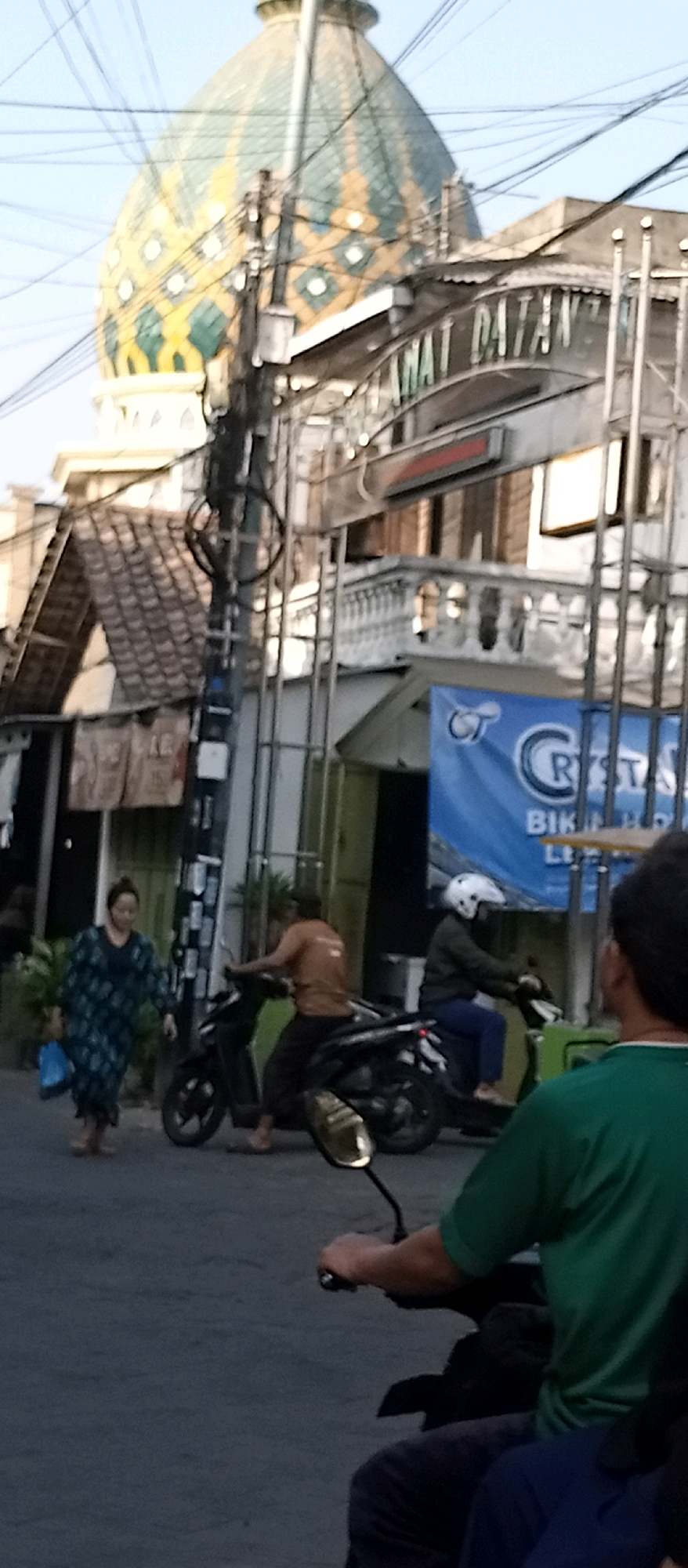 The image shows a street scene in Wadungasri, Indonesia. In the background, a distinctive dome building with green and yellow patterned tiles is visible, likely part of a mosque or similar structure. In front of it, a street sign arches over the road with text that appears to say "SELAMAT DATANG" which translates to "WELCOME" in Indonesian. A woman in a patterned blue and green dress walks on the left side of the street carrying a blue plastic bag. Two men on motorcycles are in the middle of the street. One man, wearing a brown shirt, is stopped on his motorcycle. Another man in a dark jacket and white helmet is riding his motorcycle past. In the foreground, the back of a person wearing a green shirt and dark pants is visible, seemingly riding a motorcycle and looking forward. The street is paved, and there are utility poles with numerous wires running across the scene. Buildings with tiled roofs and balconies line the street. A blue banner with white text, possibly advertising a business, is hanging from a pole on the right side. The overall lighting suggests it is daytime, possibly late afternoon due to the angle of the light.