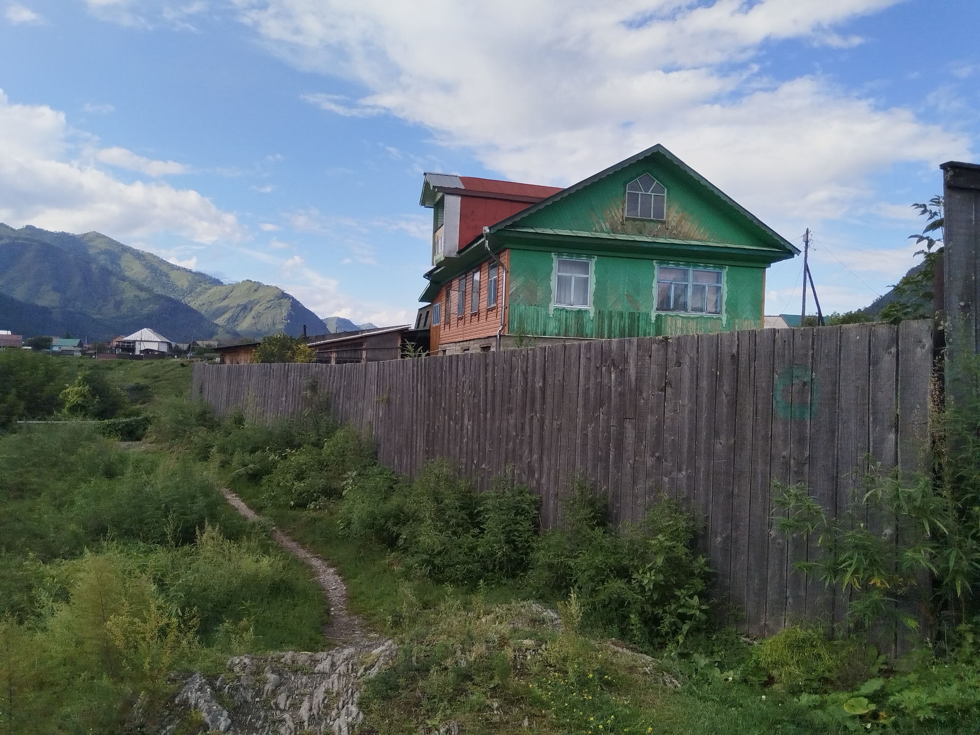 The image depicts a rural scene with a wooden fence in the foreground, a two-story green and orange house behind it, and mountains in the background under a blue sky with white clouds. A narrow dirt path winds through overgrown vegetation on the left side of the fence. The house has a red roof and several windows. A wooden pole with wires is visible to the right of the house. The overall environment appears to be a village or countryside setting, possibly in a mountainous region.