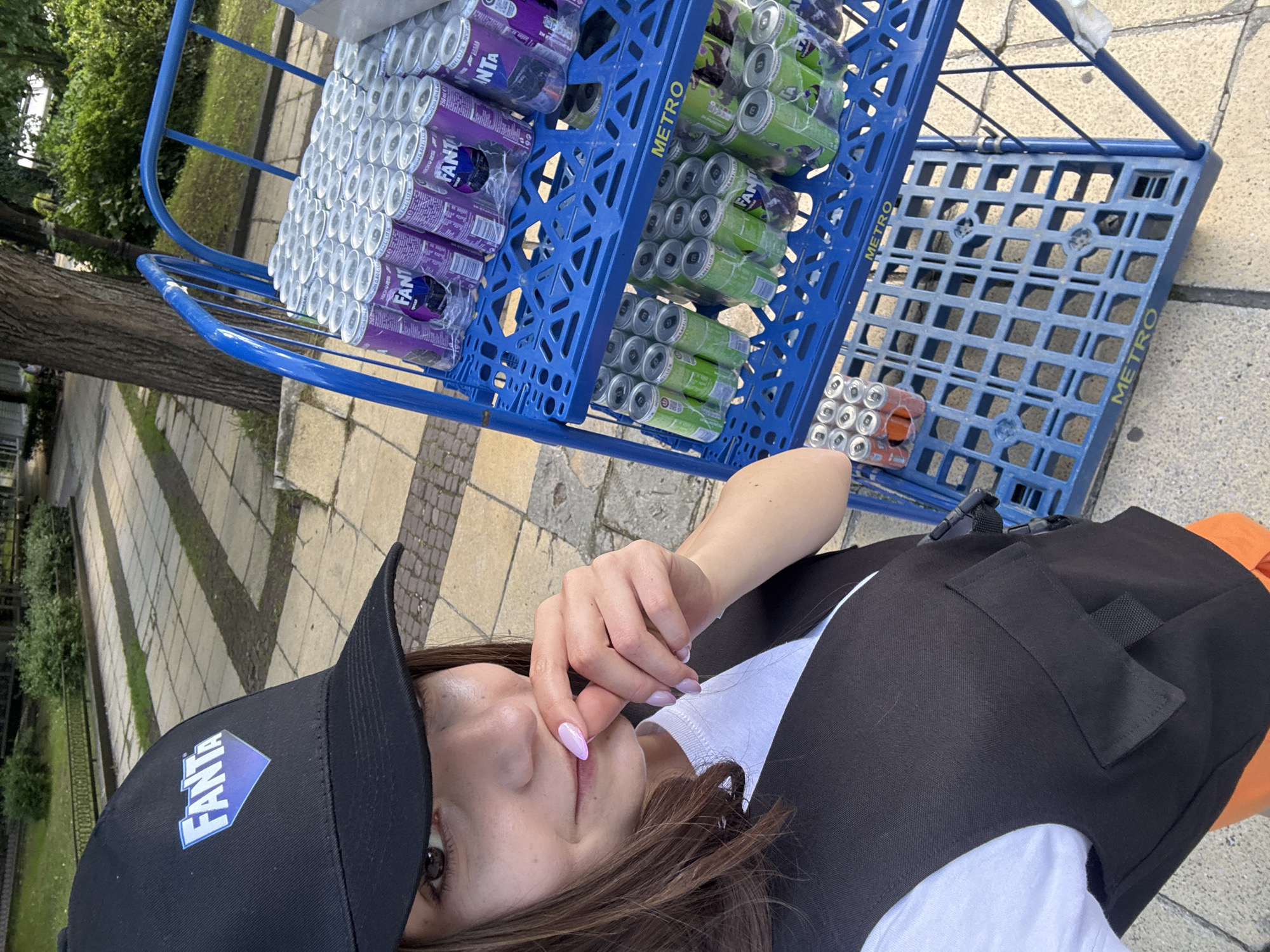 A woman wearing a black "FANTA" baseball cap, a white t-shirt, and a black vest stands next to a blue Metro brand shopping cart containing canned Fanta soft drinks in grape and green apple flavors, and orange Fanta. The scene is located outside on a sidewalk paved with rectangular stones in Varna, Bulgaria. A tree is visible to the left of the cart, and greenery lines the path.