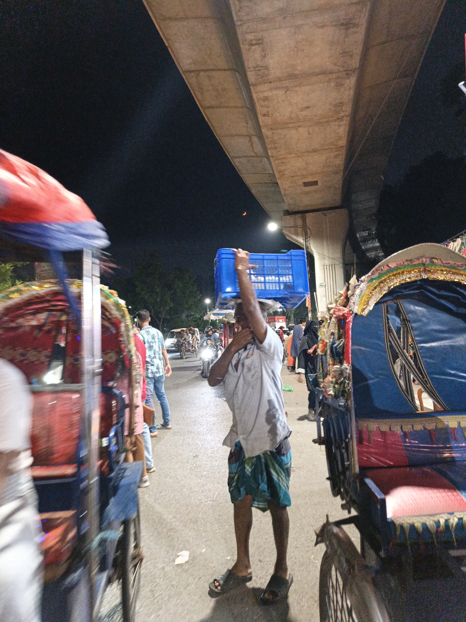 An outdoor, low-light, nighttime shot depicts a street scene beneath a concrete overpass. A man in the foreground, facing away from the camera, carries a blue plastic crate on his head. He wears a light-colored, short-sleeved shirt and a green and blue plaid lungi. To his left, a brightly decorated rickshaw is visible. In the background, several other rickshaws and motorcycles are present, along with other pedestrians. The overpass casts a shadow over the scene. The overall atmosphere is one of urban activity at night.