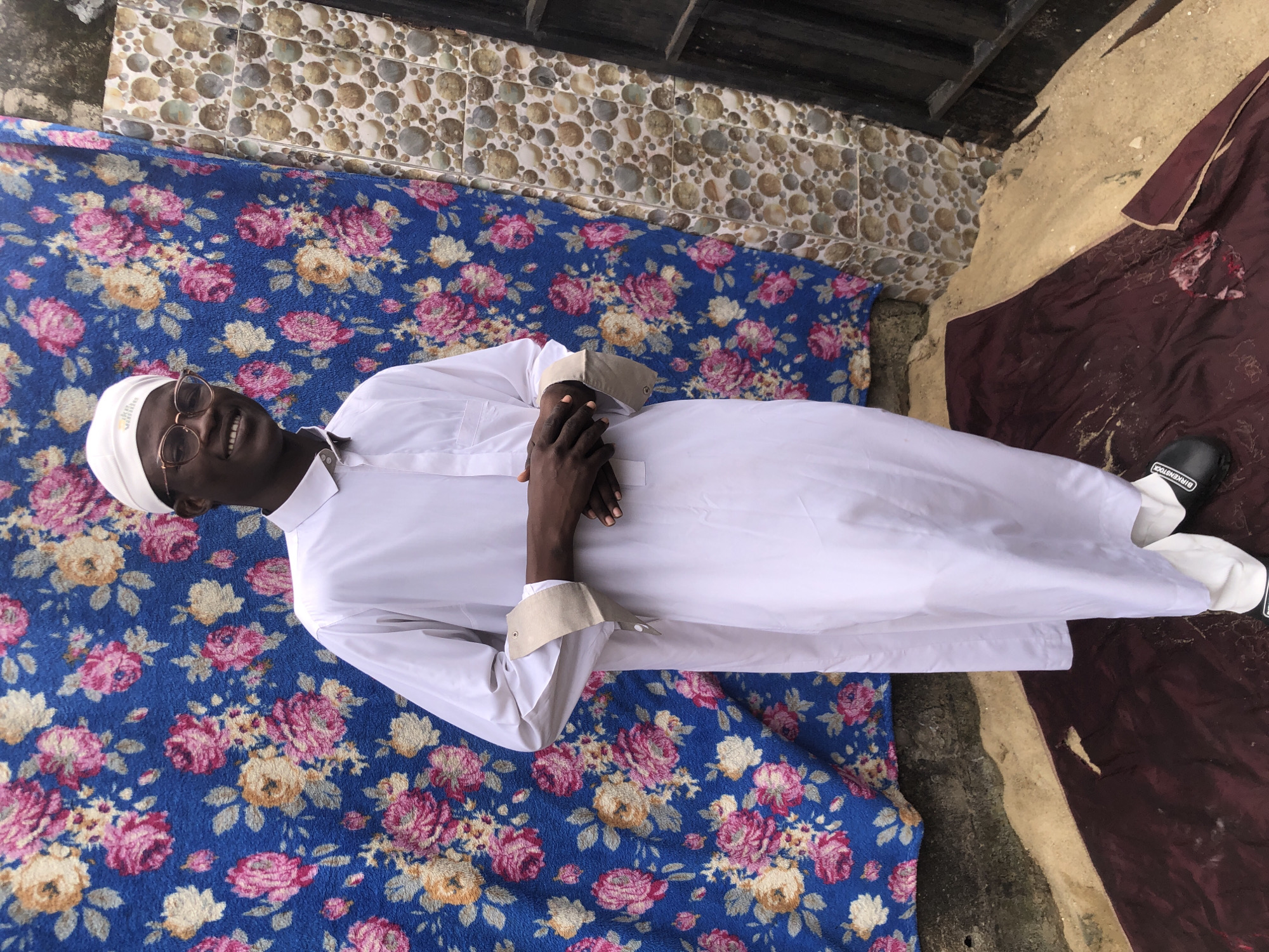 A young Black male stands in front of a blue floral fabric backdrop with a tiled wall to his right. He is wearing a white thobe and a white cap. His hands are clasped in front of him. He is smiling and wearing glasses. The ground in front of him is covered with a dark red cloth and sand. The scene appears to be outdoors, possibly in a courtyard or alleyway in Ado, Nigeria. The background suggests a modest urban environment.