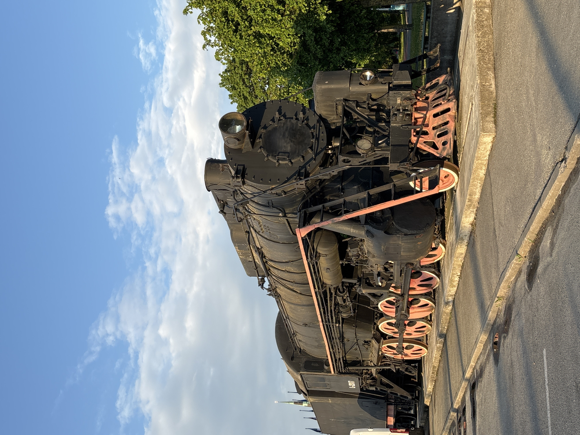 A large, black steam locomotive is prominently featured, parked outdoors under a clear blue sky with scattered white clouds. The train, an imposing piece of machinery, is viewed from its front-right side. Its main body is dark, contrasting with the vibrant reddish-orange color of its large driving wheels and a horizontal bar running along its side. Notable details of the locomotive include its substantial smokebox, a bright headlamp on the front, and intricate piping along its boiler. On the side of the train, the identification number "L-2317" is clearly visible.
The locomotive rests next to a concrete curb, beyond which lies an asphalt surface, possibly a pathway or road. The lighting suggests either early morning or late afternoon, as warm sunlight illuminates parts of the train and casts long shadows on the ground. Lush green trees form a dense backdrop behind the locomotive, indicating a park or green area setting. No people are present, and the scene depicts a static display or monument in Tallinn, Estonia.
