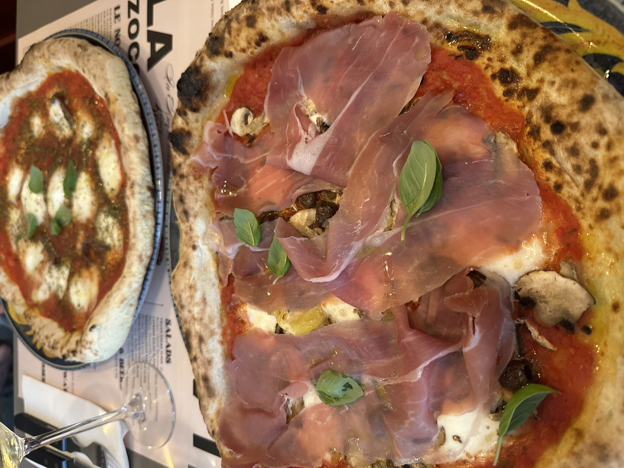 This is a close-up, overhead shot of two pizzas on a table, with a wine glass in the upper left corner. The pizza in the foreground is a large, round Neapolitan-style pizza topped with tomato sauce, mozzarella, thinly sliced prosciutto, mushrooms, and fresh basil leaves. The crust is charred in places, indicating it was cooked in a very hot oven. The pizza in the background, partially visible, is smaller and appears to be a Margherita pizza with tomato sauce, mozzarella, and basil. Both pizzas are on what appear to be paper placemats or menus. The overall setting suggests a casual dining experience, likely a pizzeria in Amsterdam, Netherlands.