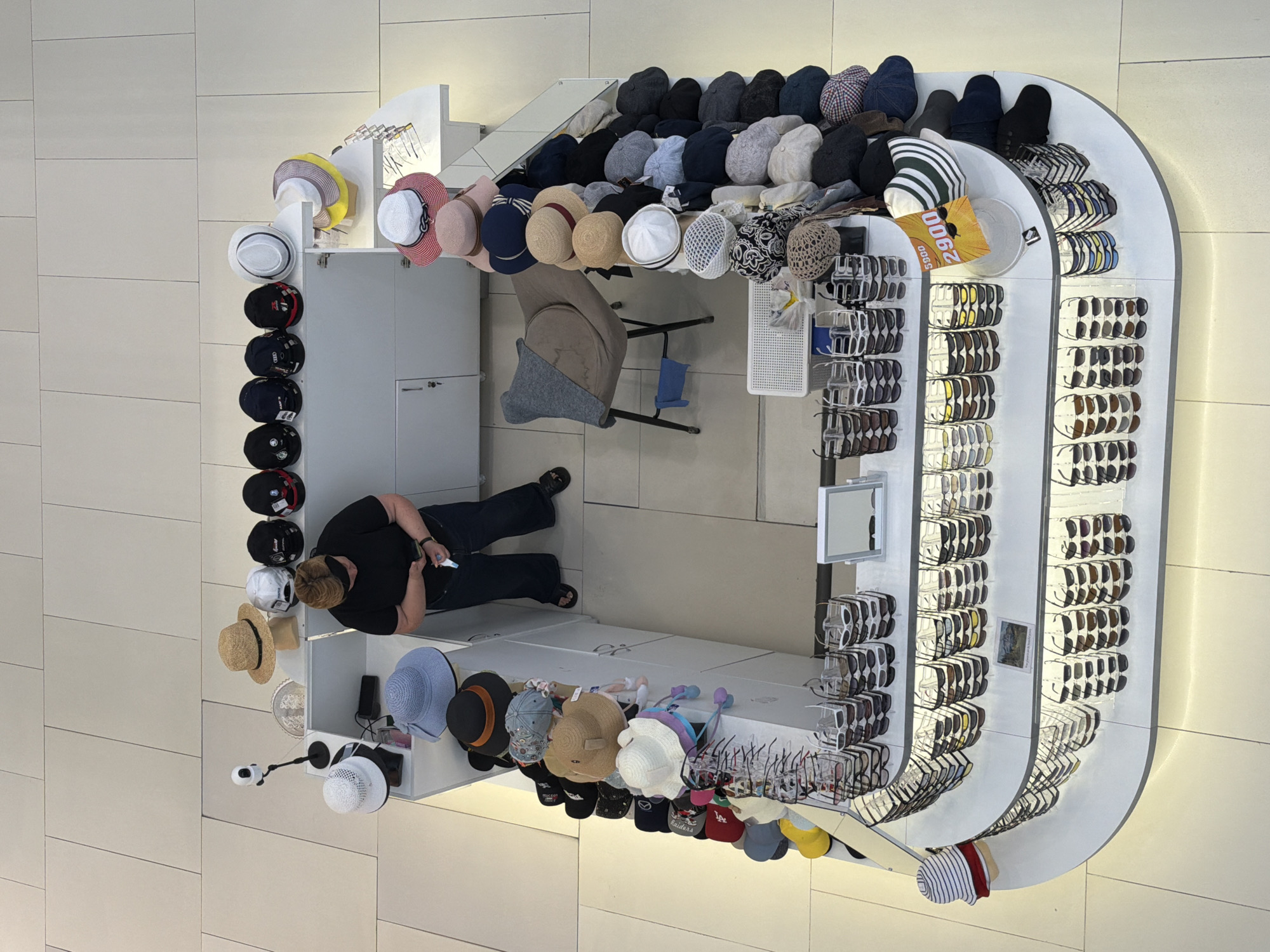The image shows a high-angle view of a retail kiosk selling hats and sunglasses. The kiosk is located in a brightly lit, modern shopping mall with a tiled floor. A woman wearing a black t-shirt and dark pants is standing behind the counter, looking at her phone. She is wearing a black blindfold around her eyes. The kiosk displays a variety of hats, including straw hats, baseball caps, and bucket hats, arranged on shelves and on a mannequin head. Sunglasses are also displayed in multiple rows on tiered shelving units. A price tag with "2900" in red text is visible on a display of hats. The overall environment suggests a commercial retail setting. There is no indication of the specific location within the city of Krasnodar, Russia.
