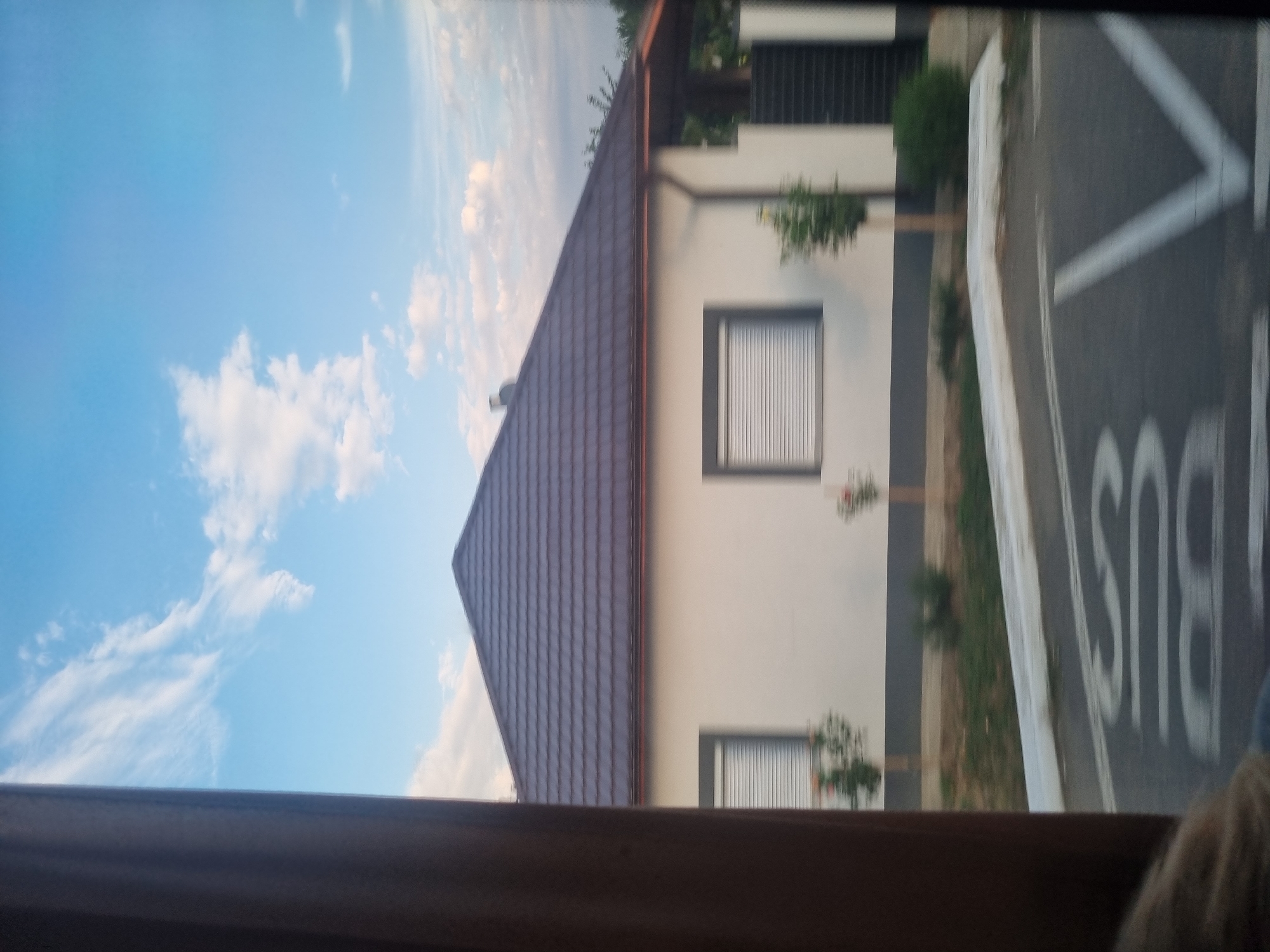 This is a photograph taken from a vehicle, looking out at a single-story house with a brown tiled roof in Gepiu, Romania. The house is white with dark trim around the windows, which have white blinds. Small trees or bushes are planted in front of the house. To the right, a dark metal fence is visible. The foreground shows a road with "BUS" painted on it in white. The sky is blue with scattered white clouds. A brown curtain or pillar is visible on the left side of the frame, obscuring part of the view.