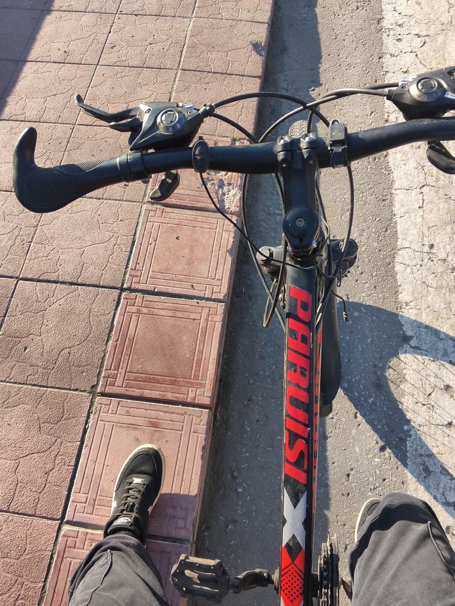 The image shows a first-person perspective of someone sitting on a bicycle. The bicycle is black with red and white lettering that reads "PARUS" and "X". The handlebars are visible, with brake levers and a gear shifter on the left side. The rider's left leg, wearing black pants and a black Nike sneaker, is positioned on the left pedal. The right leg is out of frame. The bicycle is positioned on a paved surface, with a tiled walkway to the left and a cracked concrete surface to the right, indicating an urban or suburban environment. The lighting suggests it is daytime, with shadows cast on the ground. The scene appears to be located in the city of Mokshan, Russia, based on the context provided.