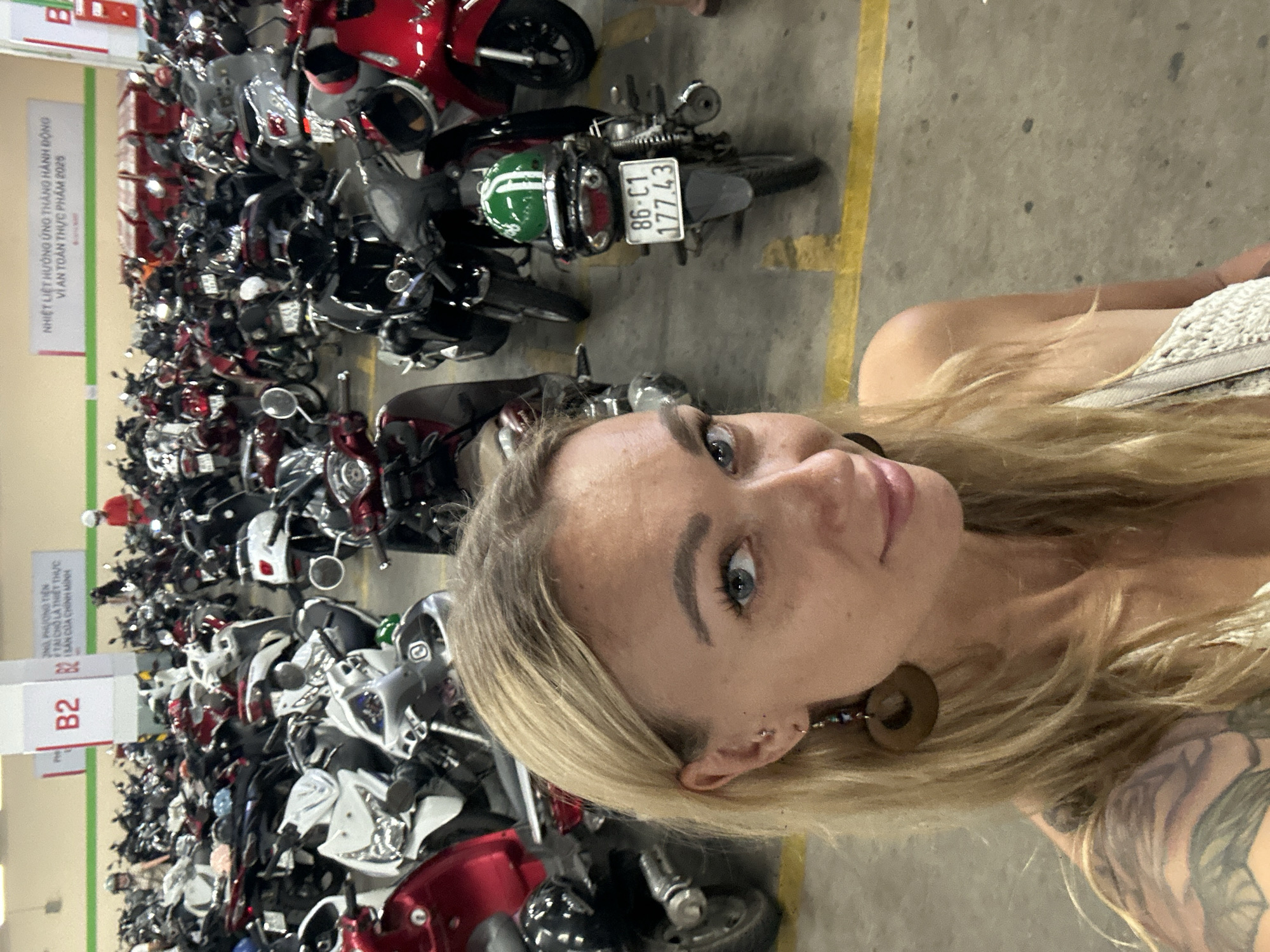 The image captures a selfie taken by a blonde-haired woman in what appears to be a large, busy motorcycle parking area. Many motorcycles of varying colors and models are parked closely together, occupying a significant portion of the frame. A sign is partially visible on the left, with a "B2" designation, with text indicating "ONG PHƯƠNG TIỆN TẠI CHỖ LÀ THIẾT THỰC". Another sign mentions "NHIỆT LIỆT HƯỞNG ỨNG THÀNG HÀNH ĐỘNG VÌ AN TOÀN THỰC PHẨM 2025." One motorcycle's license plate reads "86-C1 177.43". The location, as discerned from the sign's text, is likely within Phu Thuy Ward, Vietnam.