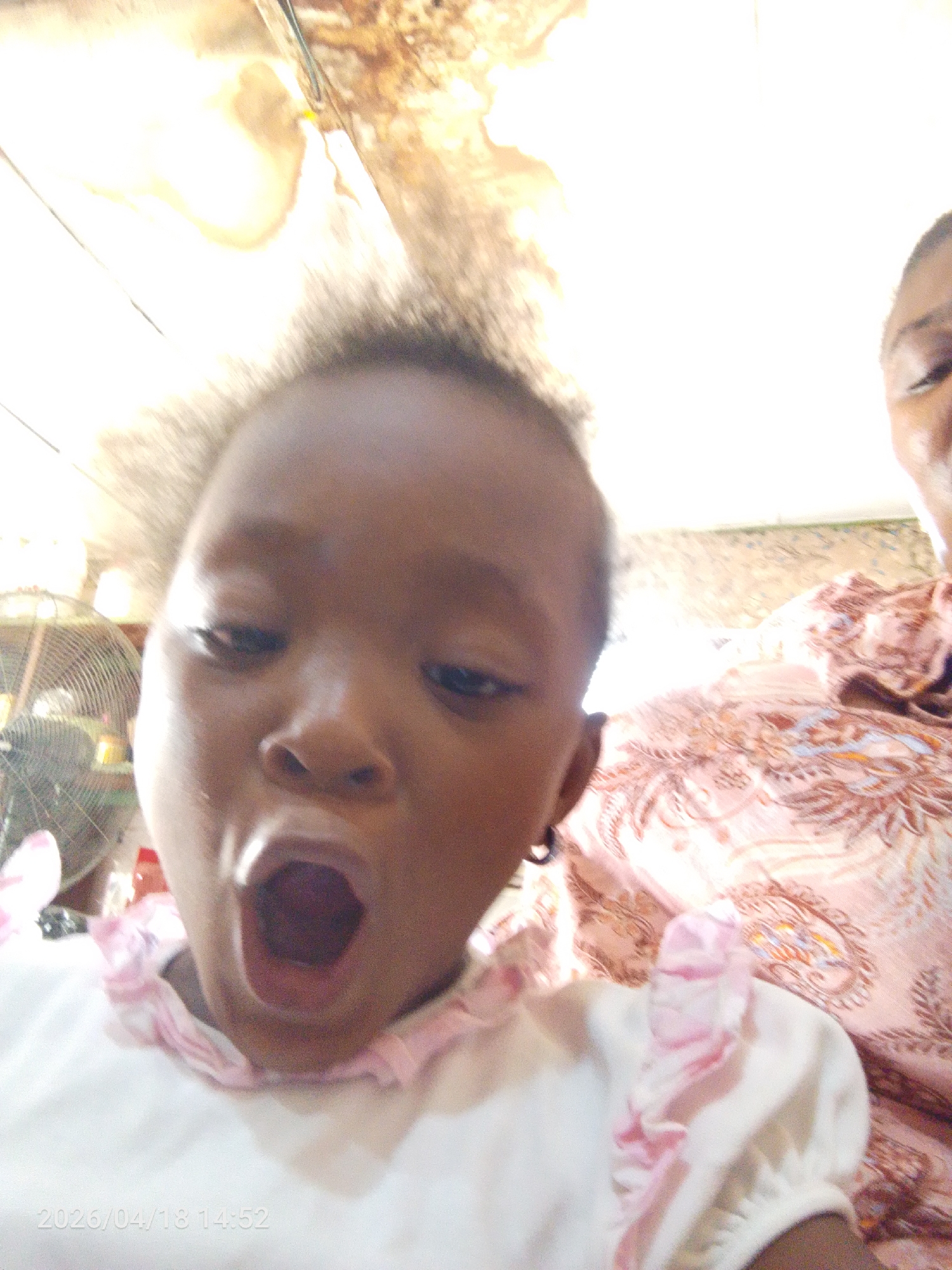 A close-up, slightly blurry photo depicts a young child with dark skin, possibly female, with their mouth wide open. The child is wearing a white shirt with pink ruffles around the collar. Part of an adult's face is visible in the upper right corner, wearing a pink and brown patterned shirt. An electric fan is visible in the background, on the left. The photo appears to have been taken indoors. The timestamp indicates April 18, 2026. The specific location within Lokoja, Nigeria is not evident from the photo.