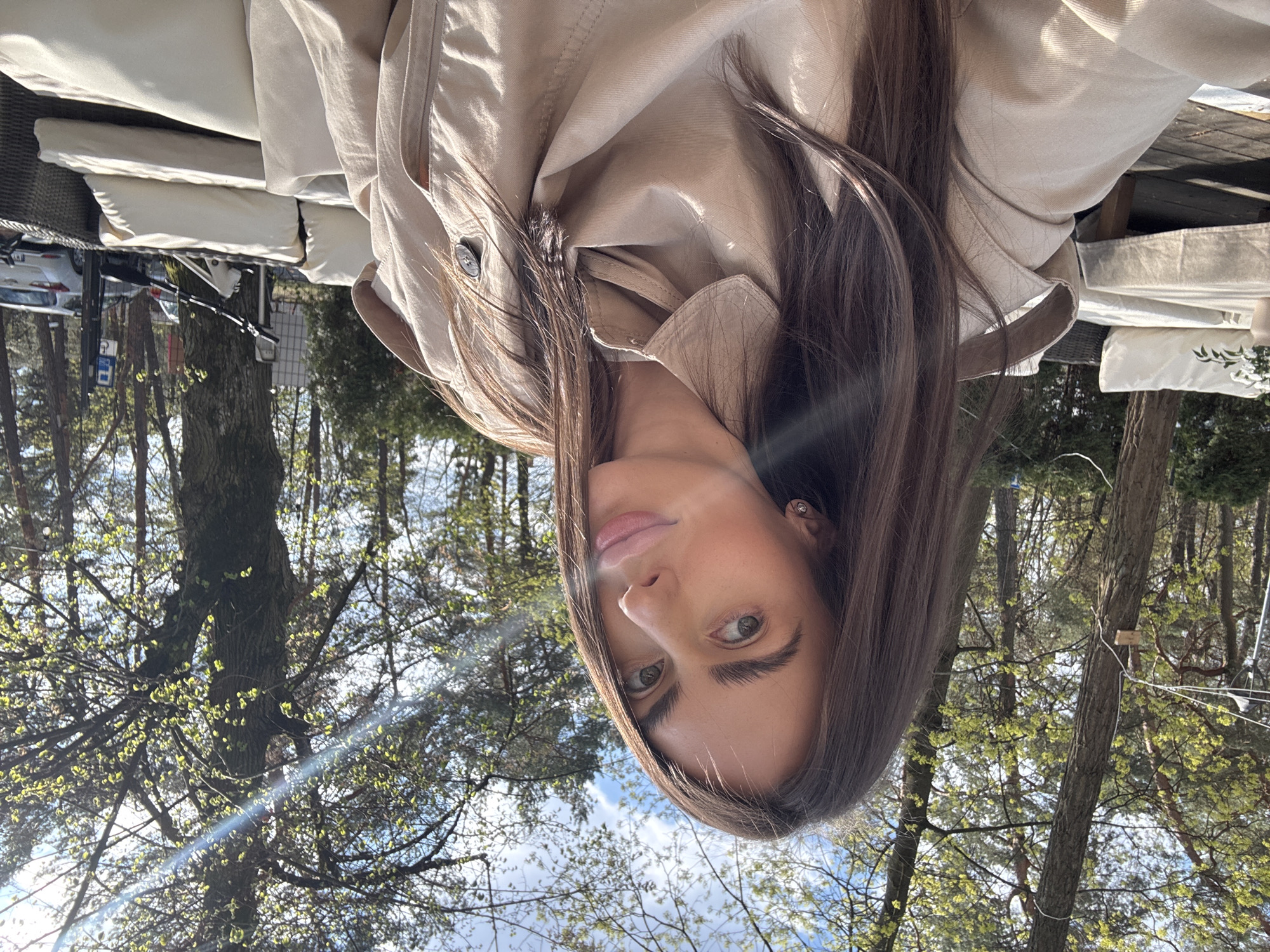 A woman is the primary subject, facing the camera. She has brown hair and is wearing a tan coat. The background shows trees and a bright sky. Based on environmental context and the presence of trees, this picture could be taken in Konstancin-Jeziorna, Poland, a town known for its parks and greenery. The woman appears to be sitting outside, possibly at a cafe or outdoor seating area. Cushions are visible behind her. The overall setting is sunny and pleasant.