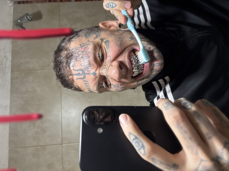 The image displays an adult male in a private bathroom setting within Buenos Aires, Argentina. The individual, characterized by extensive dark tattoos covering his face and head—including a prominent trident symbol centrally on the forehead, "MMAM" on the left temple, and "DON'T LOOK THE CHUPACABRA" on the right temple—is engaged in a self-portrait. His short, dark hair is visible, along with significant earlobe gauges. He is clad in a black tracksuit jacket featuring white stripes on the shoulders. With his left hand, he holds a black smartphone, oriented towards himself to capture the image. His right hand holds a light blue toothbrush with white bristles, which is applying friction to a set of silver-colored grillz covering his upper and lower teeth. The male's facial expression is a contorted grimace, with eyes partially closed and mouth open, revealing the grillz and toothbrush. The immediate background consists of light brown tiled walls. In the upper right quadrant, a chrome shower head is visible. Out-of-focus red objects, consistent with hanging toothbrushes or similar bathroom accessories, are present near the top edge of the frame.