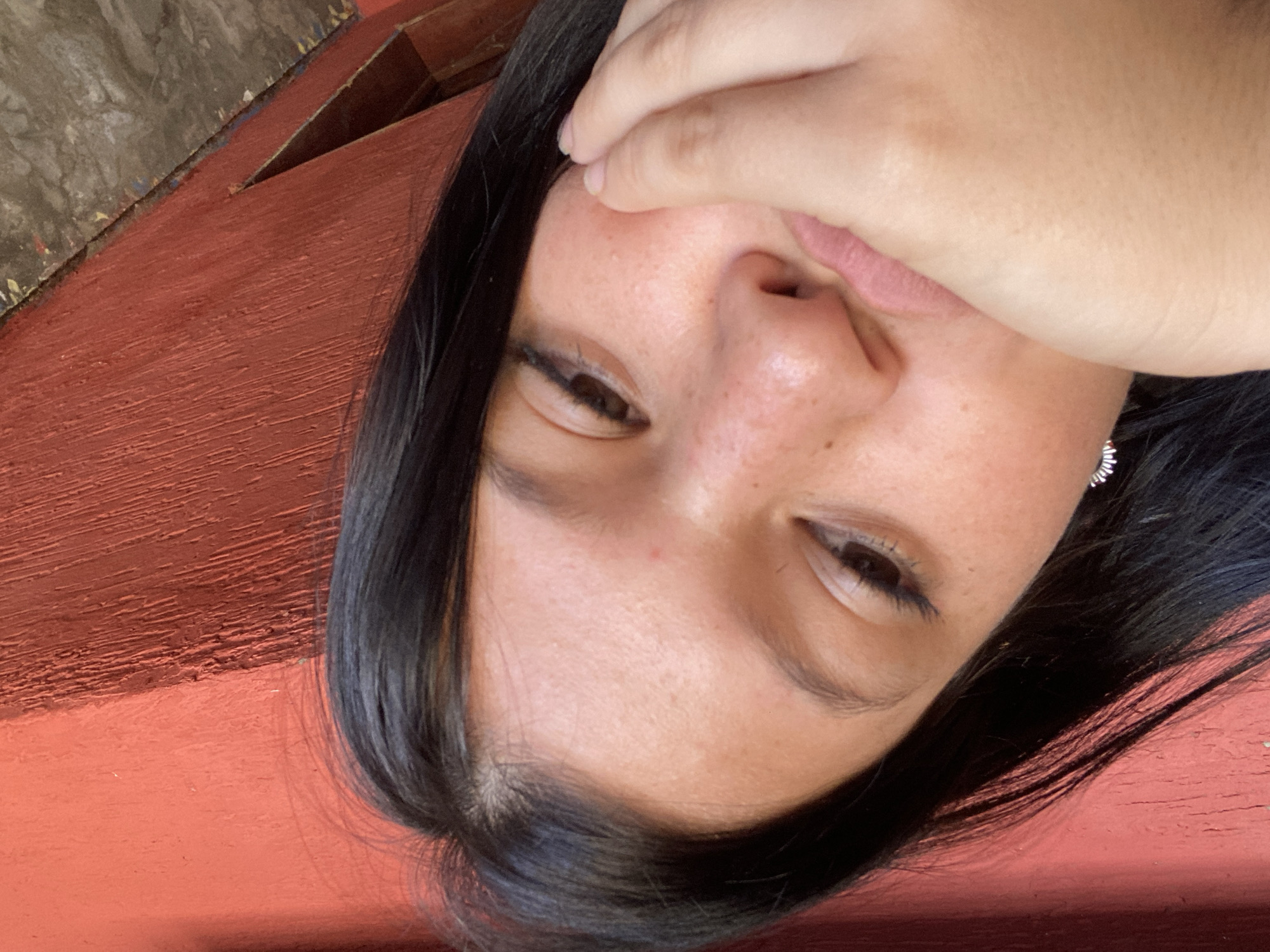 The image is a close-up shot of a woman's face, rotated. She has dark hair, fair skin, and is partially covering her face with her hand. Her eyes are partially closed. An earring is visible on her right ear. The background appears to be a reddish-brown wooden surface. The image does not contain enough information to determine its location in Juan Griego, Venezuela.