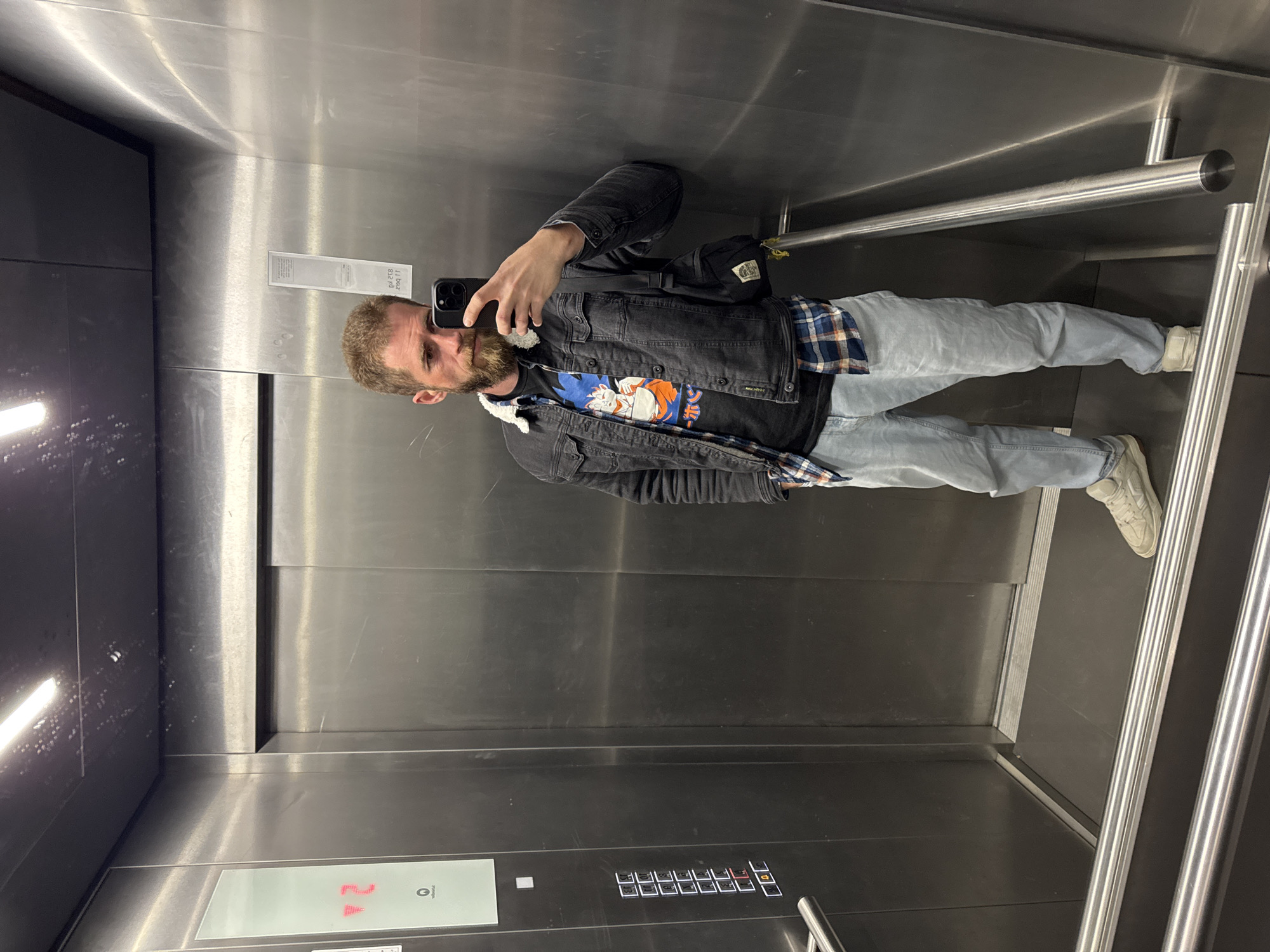 A man is taking a selfie in an elevator with stainless steel interior walls. He is wearing a denim jacket, a t-shirt with a graphic print, light blue jeans, and white sneakers. He has a beard and appears to be of Caucasian descent. The elevator has handrails and a panel with buttons and a floor indicator showing "24." The location in the city of Budva, Montenegro is not discernible from the image.