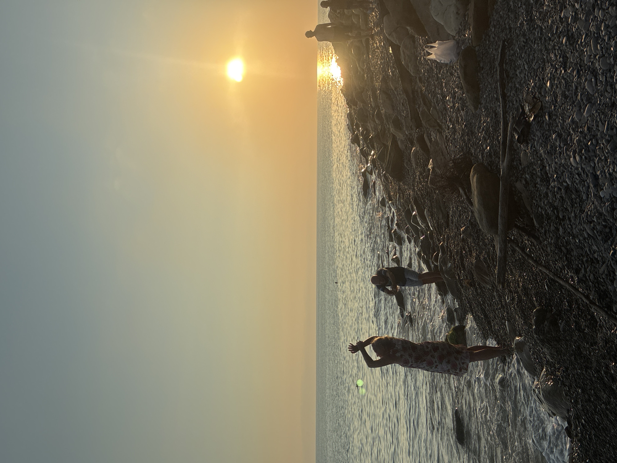 The image depicts a coastal scene at sunset, likely on the Black Sea in Sochi, Russia. The sun, a bright orb, is low on the horizon, casting a warm, golden glow across the sky and water. The water is calm, with gentle waves lapping at the rocky shore.
In the foreground, a person in a patterned dress stands with their arms raised, silhouetted against the sunset. To their right, another person is partially visible, appearing to be taking a photo or video. Further along the shore, two more figures are silhouetted in the distance, walking towards the setting sun.
The shore is covered in smooth, dark pebbles and larger rocks, with some driftwood scattered about. A white plastic bag is visible on the ground amongst the rocks. The overall impression is one of a peaceful evening by the sea.