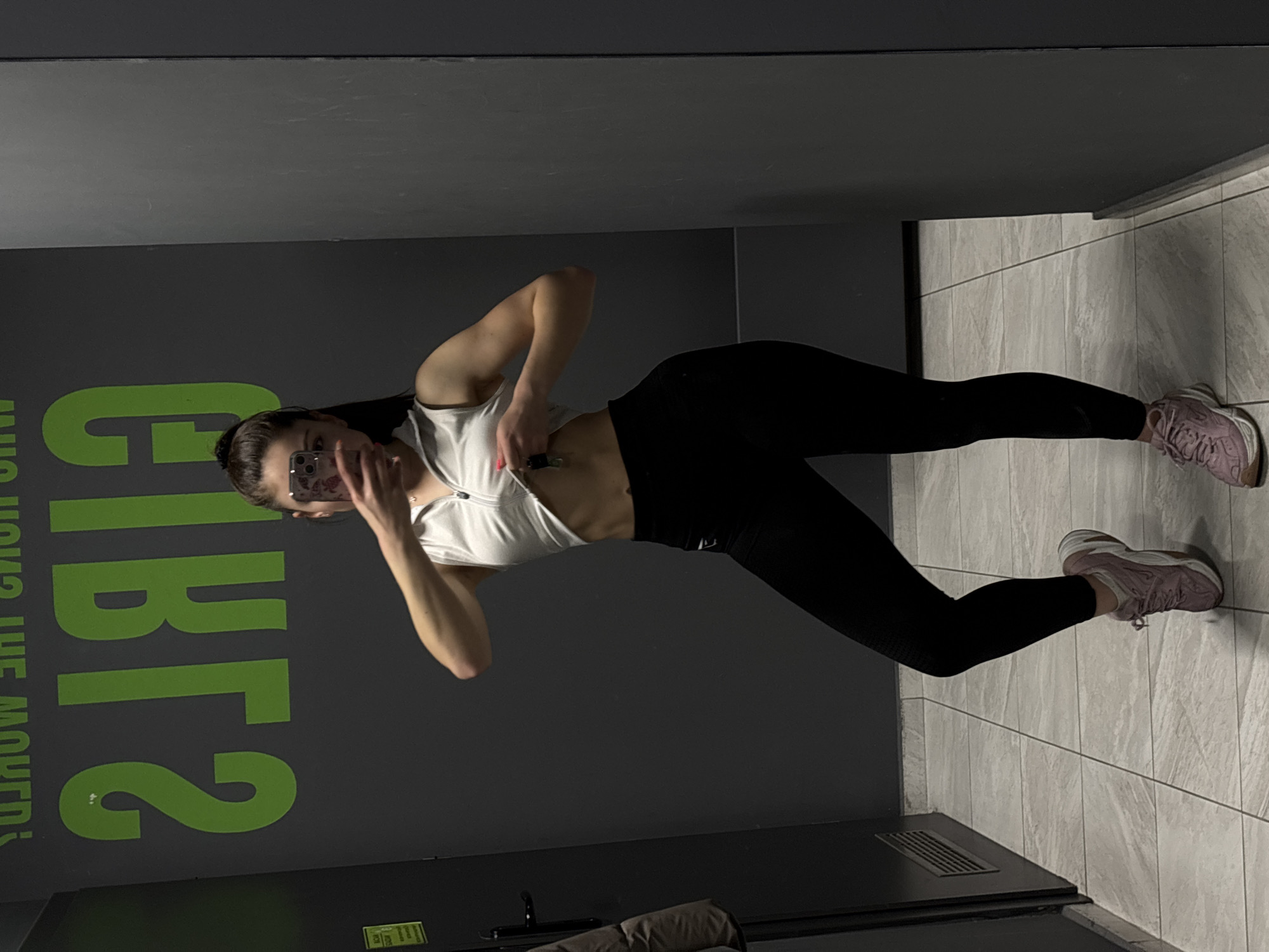 A young woman is captured in a mirror selfie, standing indoors in what appears to be a gym or fitness facility. The image is presented rotated 90 degrees counter-clockwise.
The subject is dressed in athletic attire, including a white crop top that reveals her midriff, black form-fitting leggings, and light-colored athletic shoes, possibly pink or lavender. Her dark hair is pulled back, and she holds a smartphone with her right hand, taking the photo and partially obscuring her face. Her left hand is near her stomach, holding a small, dark object, possibly a lighter or vape device, tucked into her top.
The setting features a large mirror reflecting a dark gray wall. The floor is composed of light gray tiles arranged in a grid pattern. A dark structure, possibly a bench or equipment with a ventilation grate, is visible towards the bottom edge of the image.
On the dark gray wall, as seen in the mirror's reflection, there is green text. Smaller text above reads "WHO MOVED THE MOUTH?". Below this, larger green characters are visible, appearing as "2TRATS" in the reflection (which would likely be "START2" if un-mirrored). The lighting is bright and artificial, typical of an indoor environment. The woman's facial expression is not clearly discernible due to the phone covering part of her face.