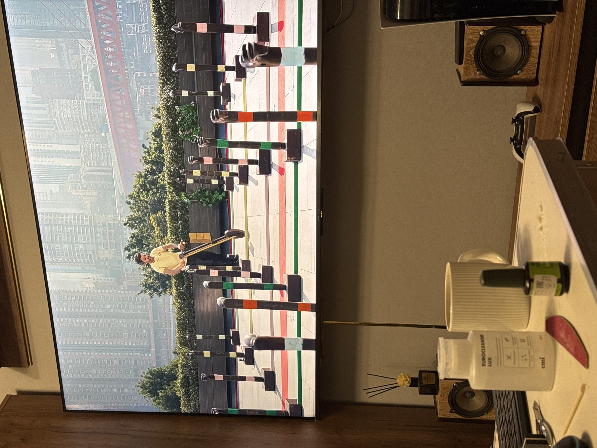 The image displays a television screen showing a man in a yellow t-shirt operating a device with wheels and a handle, navigating through a course marked by poles with colored bands. The background on the television features a cityscape with tall buildings and a red bridge. In the foreground, on a desk, are several items including a bottle labeled "EUROCLEANSER LUX," a ribbed mug, a bottle of green nail polish, a nail file, a wooden stick, a keyboard, a speaker, a PlayStation controller, and a gaming console. A diffuser with reeds and a flower is also visible on the desk. The overall environment appears to be an indoor setting, possibly a living room or office. There is no information in the image to suggest the location is Mikhaylovsk, Russia.