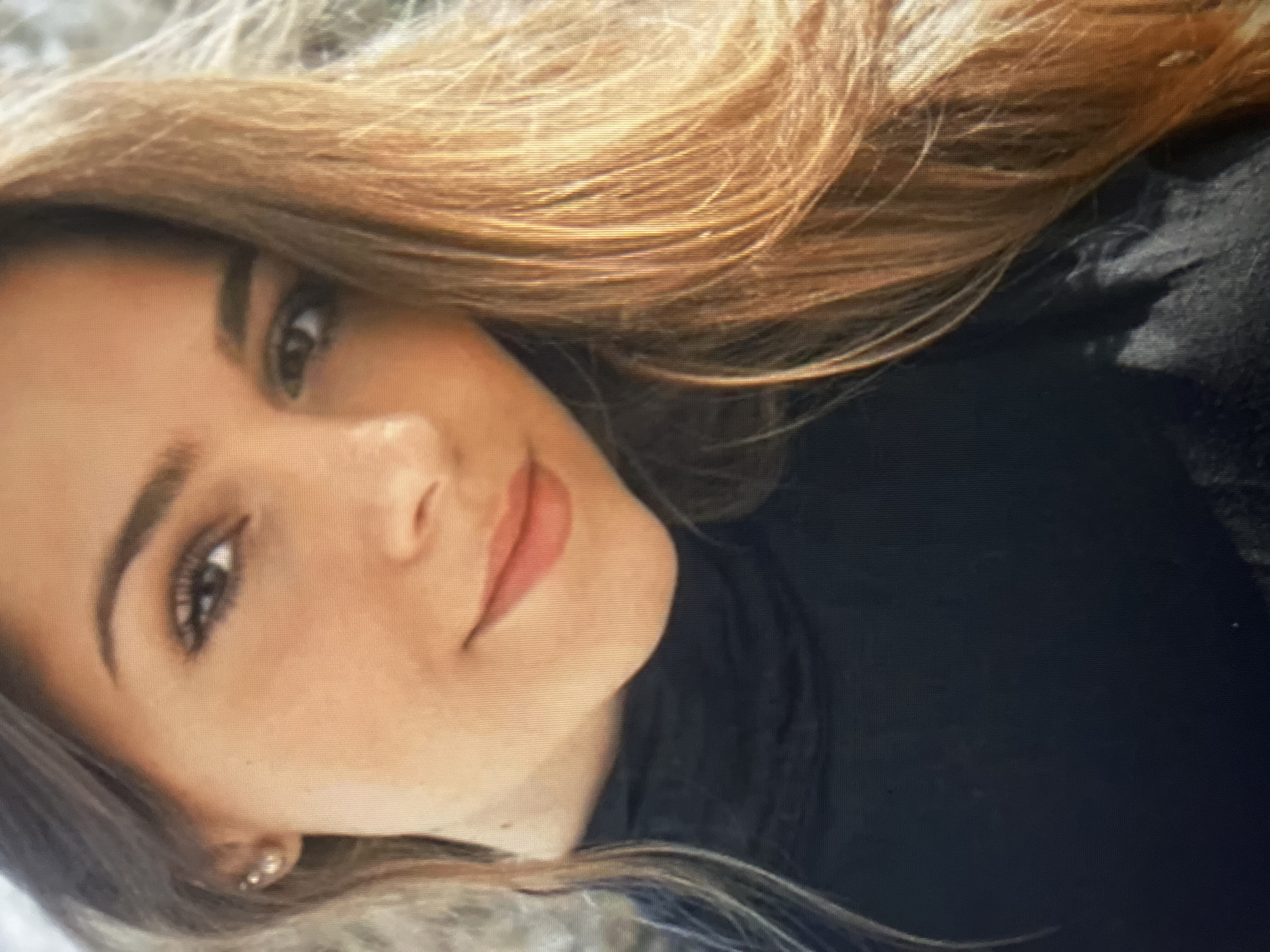 A close-up, medium-shot photograph shows the upper body of a young woman with long, light brown hair, fair skin, and a light application of makeup. She is wearing a black, high-necked garment. The background is blurred and appears to be outdoors with foliage. The lighting suggests daylight. No specific location within Qarağandı city administration, Kazakhstan, can be identified from the image.