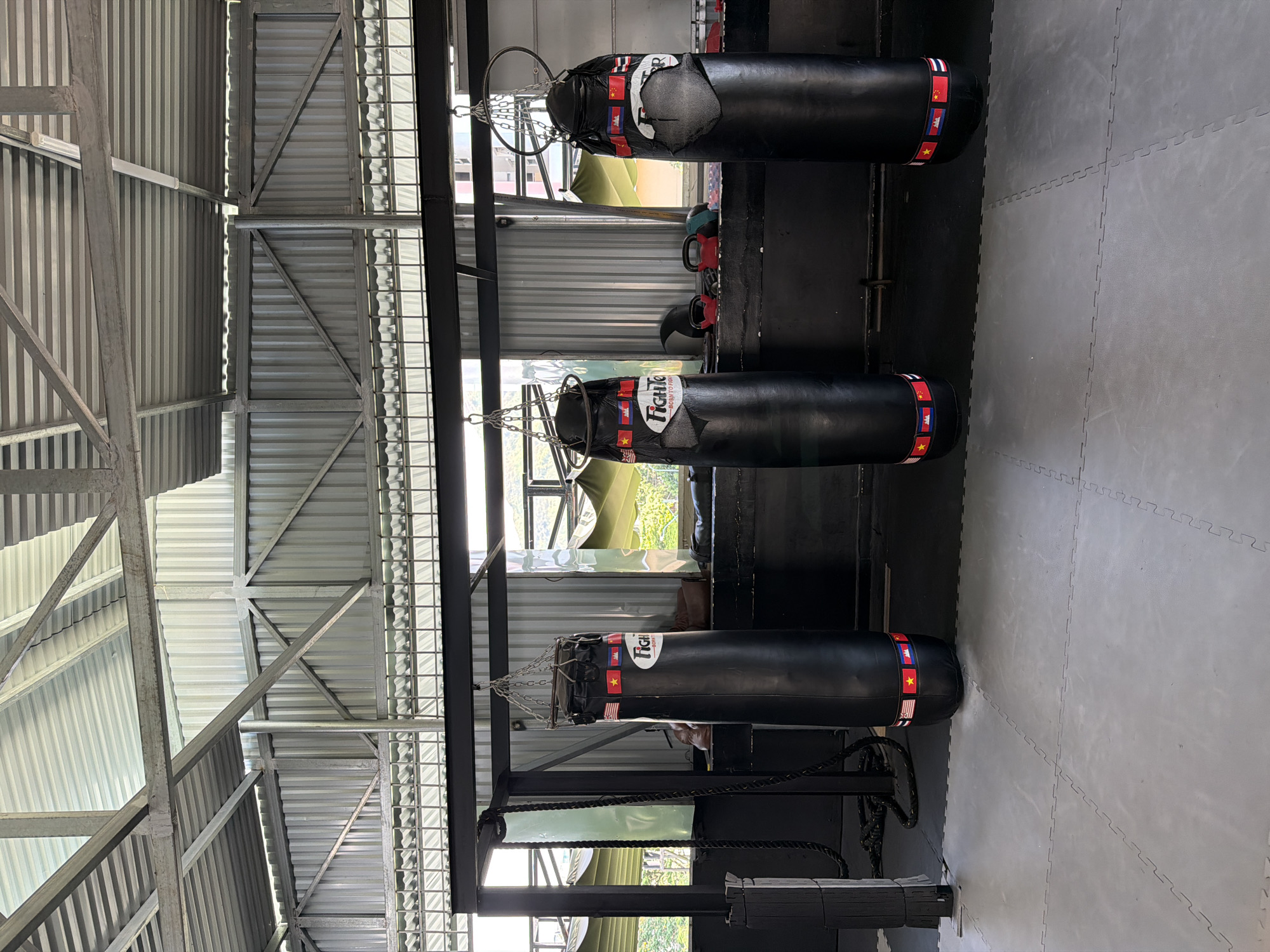 This is a mid-shot, eye-level photograph of three black punching bags hanging from a metal frame in a gym. The punching bags are lined up horizontally in the center of the frame, with the left one slightly closer to the viewer. Each punching bag has a band of international flags wrapped around its midsection. The gym has a corrugated metal roof and walls, with windows that let in natural light. The floor is covered with grey interlocking foam mats. The overall environment suggests a martial arts or boxing training facility. The scene is static with no discernible action or interaction. The lighting is natural and appears to be from daytime. The setting is indoors, likely a gym or training space in Bac Nha Trang Ward, Vietnam.