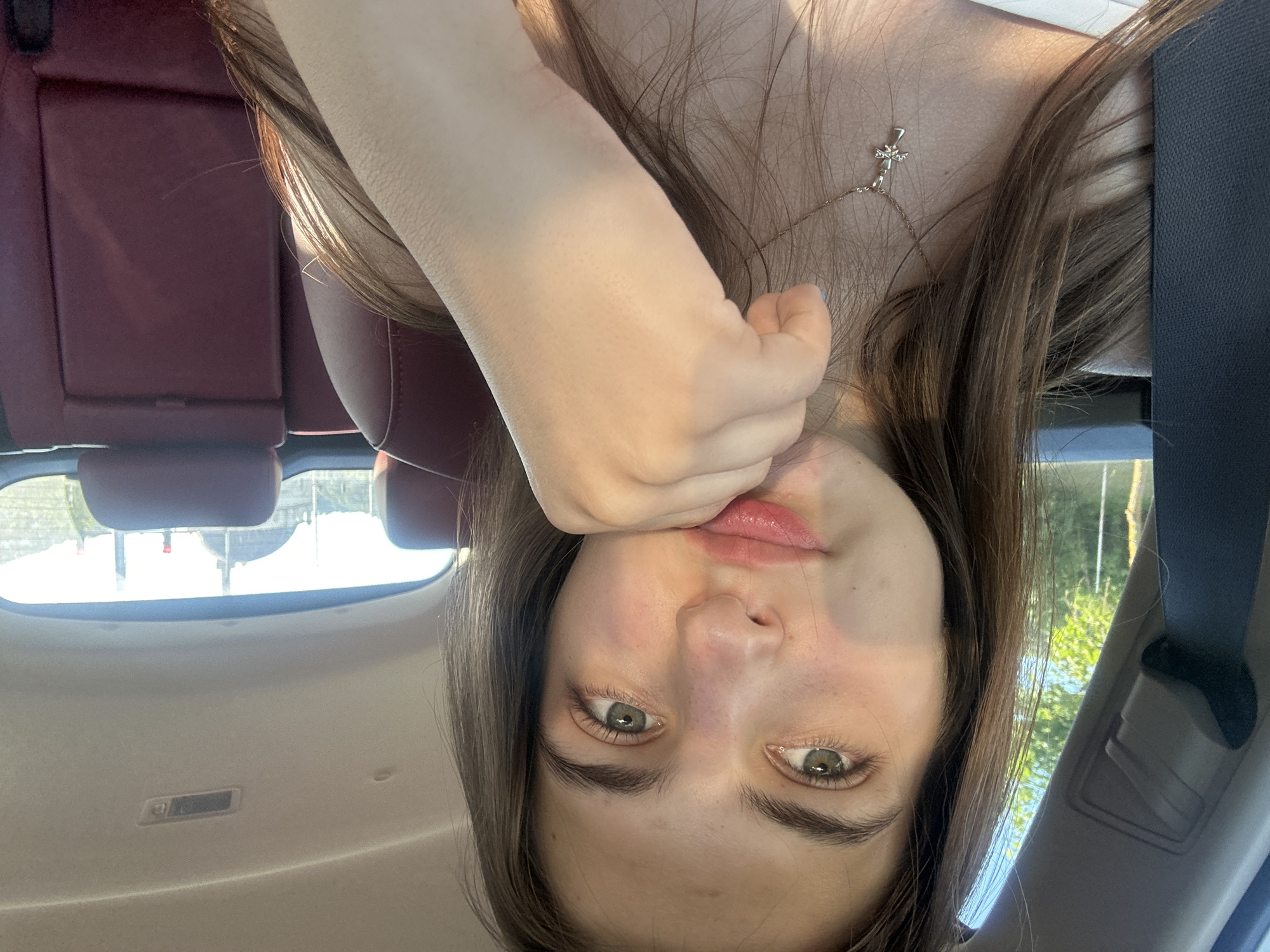 A young woman with long, brown hair is taking a selfie inside a car. She is wearing a white top and a silver cross necklace. Her right hand is resting on her chin. The background shows the interior of the car, including red leather seats and a view of trees and a building through the back window. The image is lit by natural sunlight. The image was taken in Shumen, Bulgaria.