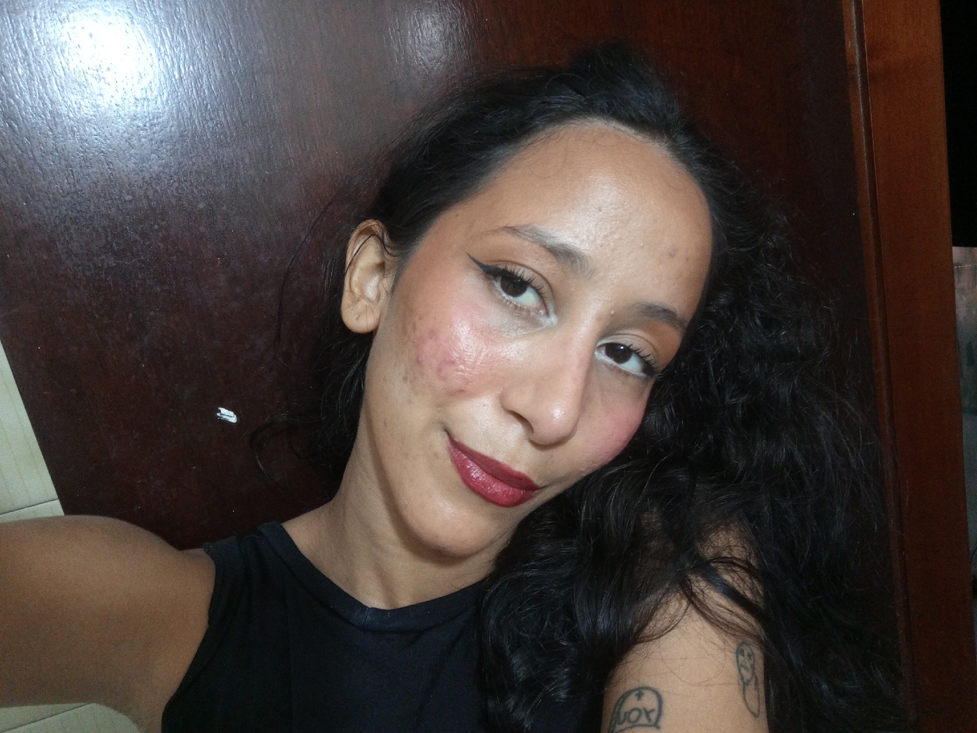 A woman is shown in a close-up selfie. She has dark hair pulled back from her face and dark curly hair falling over her right shoulder. She is wearing a black tank top. The background appears to be a dark wooden surface. The woman has dark eyes, fair skin, and is wearing makeup, including winged eyeliner and red lipstick. A tattoo is visible on her upper left arm. The specific location in the city of Porlamar, Venezuela is not identifiable from the image.