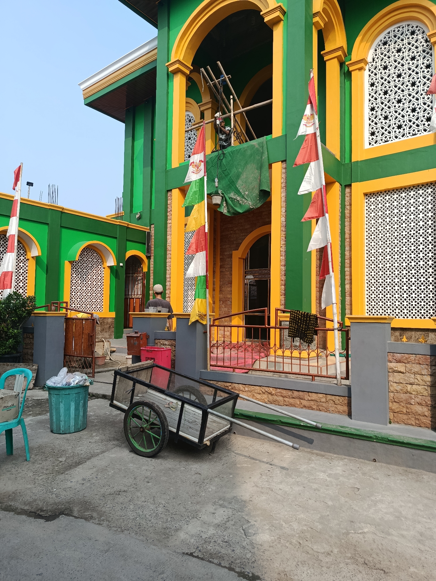 A mosque building in Balaraja, Indonesia, is pictured with a facade of green and yellow. The building features arched windows with intricate white latticework and is decorated with several Indonesian flags, which are red and white. In front of the mosque, a person wearing a white cap stands near an entrance. A small cart with wheels, a green trash can filled with refuse, and a turquoise chair are situated on the concrete ground in the foreground. The sky is clear and blue.