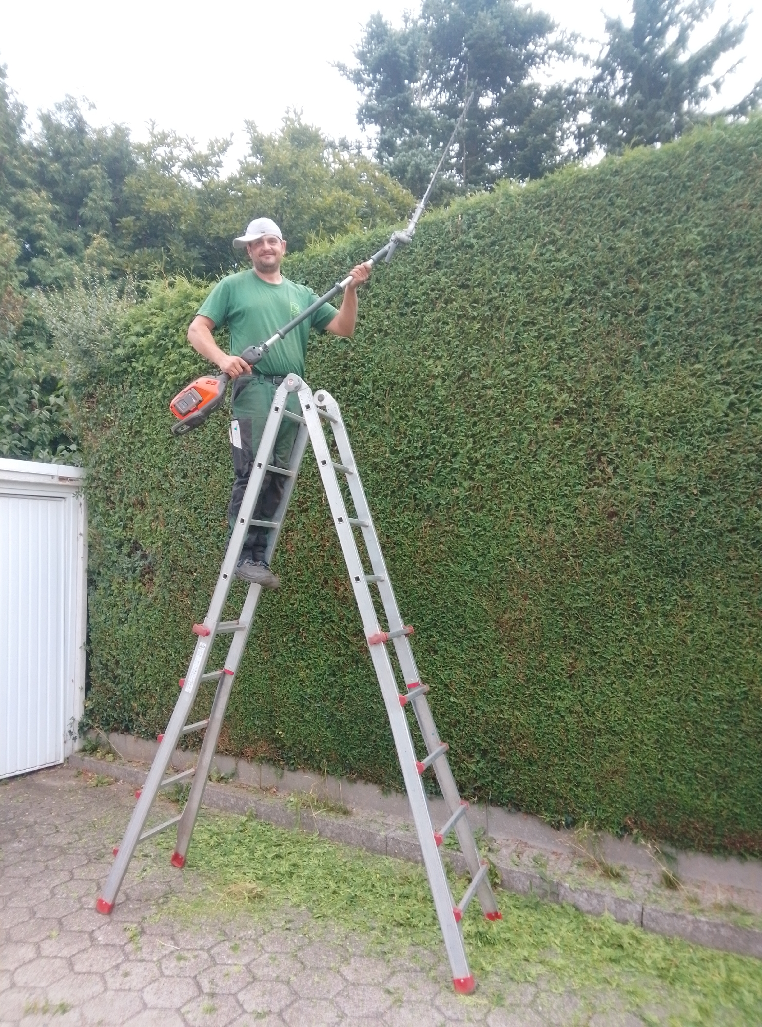 A man in a green t-shirt and green overalls stands on a silver ladder, holding an orange and black hedge trimmer. The hedge trimmer has a long pole. He is positioned in front of a tall, green hedge. The ladder is set up on a grey paved area with hexagonal tiles. There are clippings of grass scattered on the ground. A white garage door is visible to the left of the scene. The image appears to be taken outdoors, likely in a residential area of Wedel, Germany, given the hedge and the presence of a garage. The man is smiling at the camera and appears to be in the process of trimming the hedge.