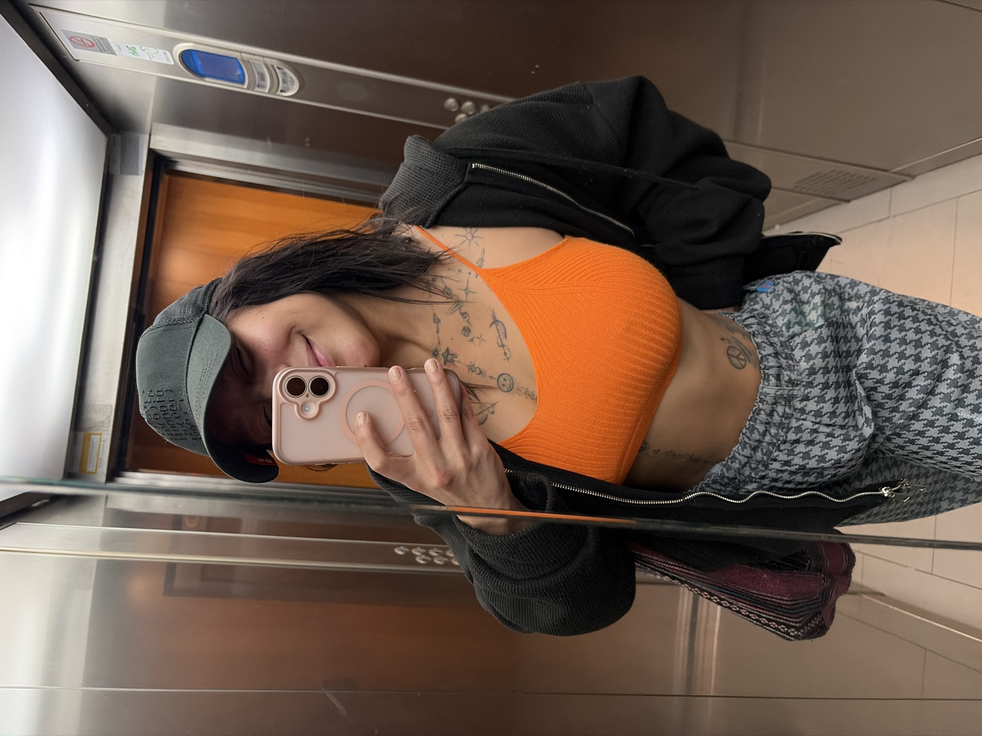 A person stands inside an elevator in L'Hospitalet De Llobregat, Spain, taking a selfie in the mirror. They are wearing a dark baseball cap, an orange ribbed tank top, a black zip-up jacket, and houndstooth pants. Visible on their skin are tattoos. The person is holding a pink cell phone. The elevator interior features brushed metal walls, a wood-paneled wall, and control panel.