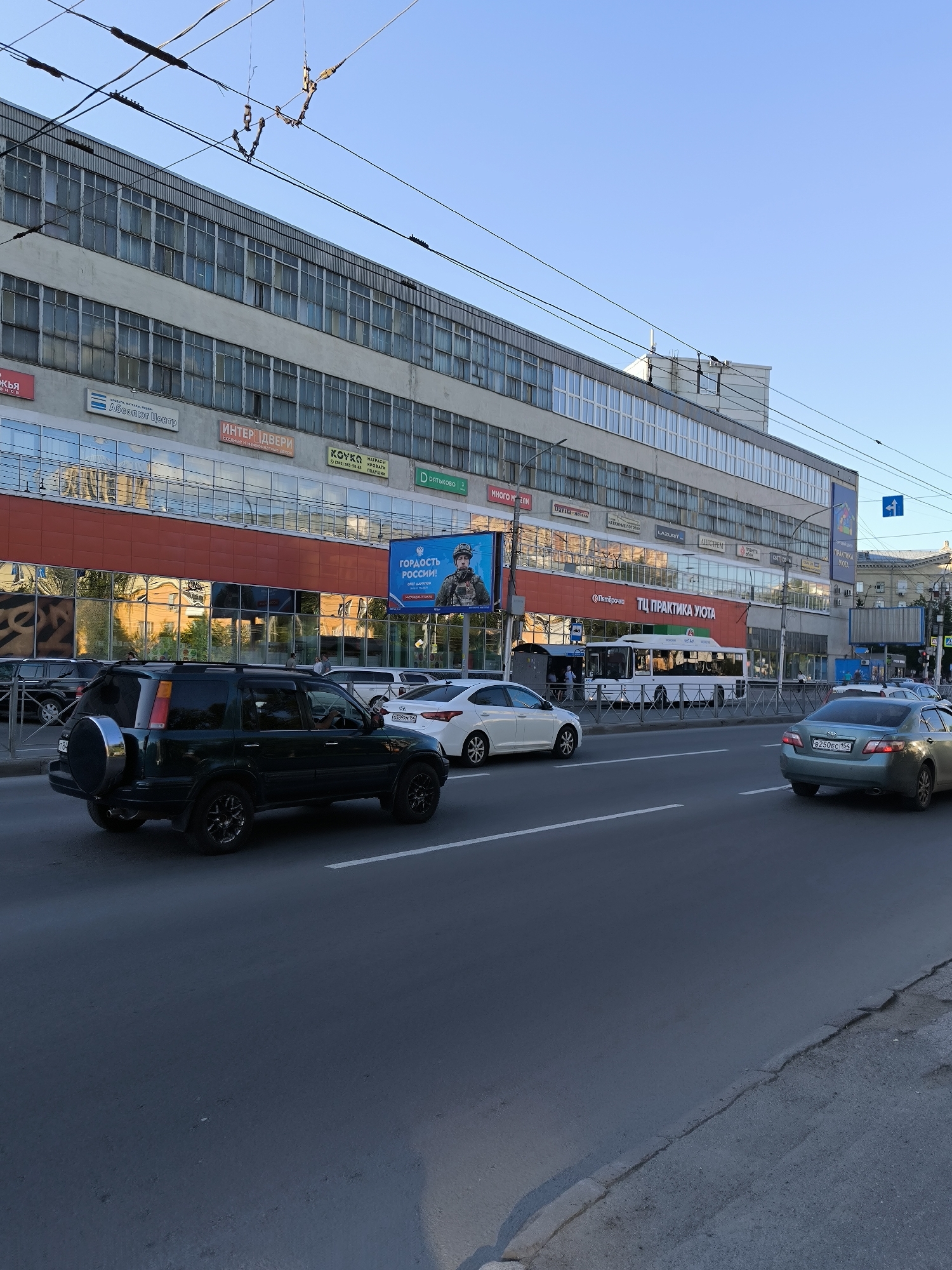 The image shows a street scene in Novosibirsk, Russia. A large, multi-story building with numerous windows lines the street. Various signs are visible on the building, including "Абсолют Центр", "ИНТЕР ДВЕРИ", "КОУКА", and "ТЦ ПРАКТИКА УЮТА". A billboard displaying a soldier and the text "ГОРДОСТЬ РОССИИ!" is also present. Several cars are on the road, including a dark green SUV, a white sedan, and a light green sedan. A white bus is also visible. Overhead power lines crisscross the sky. The scene appears to be a commercial or mixed-use area in the city.
