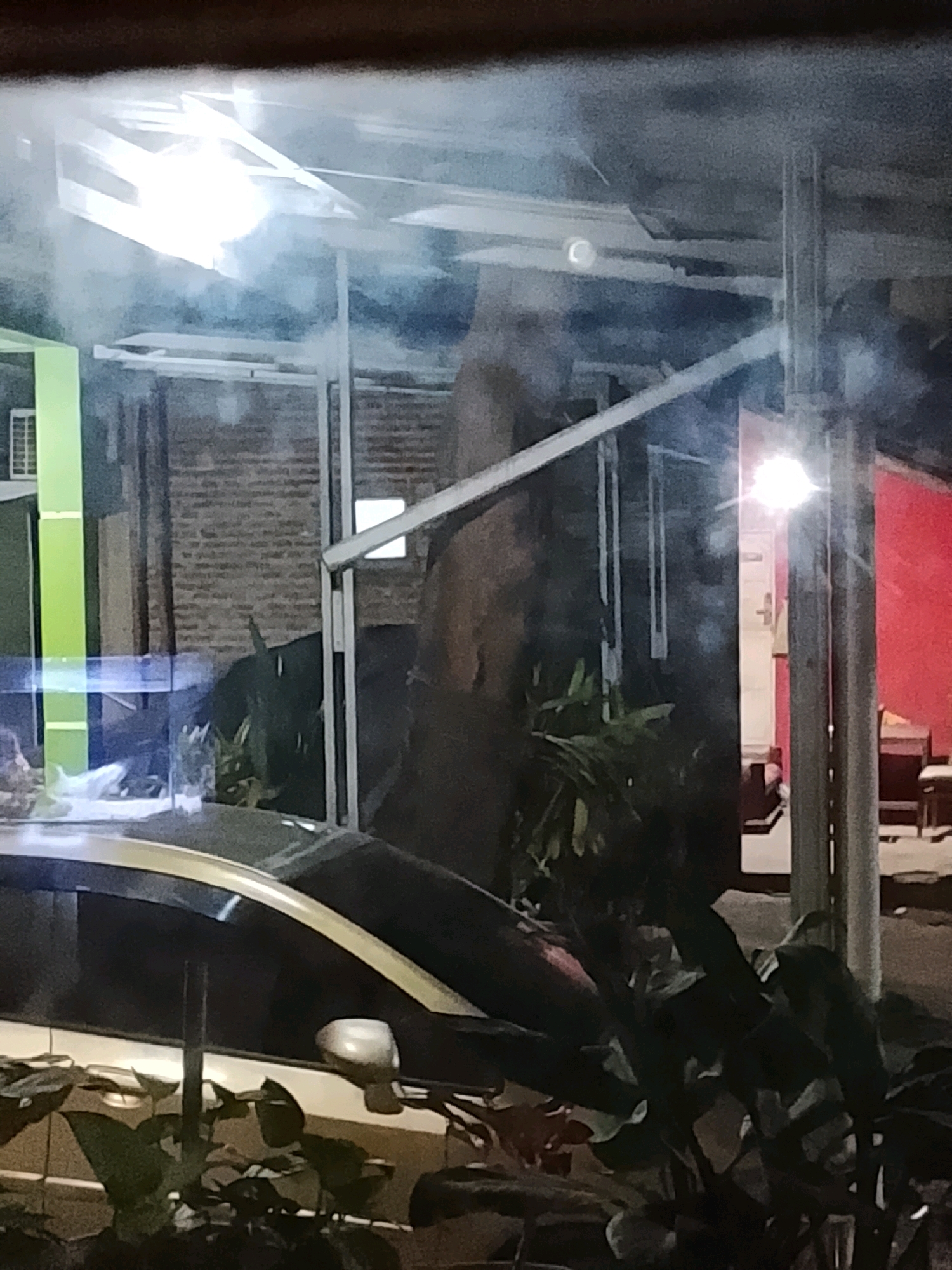 The image shows a nighttime view of an outdoor area, possibly a parking lot or a walkway, with a car in the foreground. The car appears to be a light-colored sedan, partially obscured by dark foliage. Overhead, a metal structure with lights is visible, casting a bright glare. In the background, a brick wall and a red-painted wall are present. There are some plants and trees in the scene. The overall lighting is dim, with strong highlights from artificial light sources. The location is identified as Ujungrusi, Indonesia.