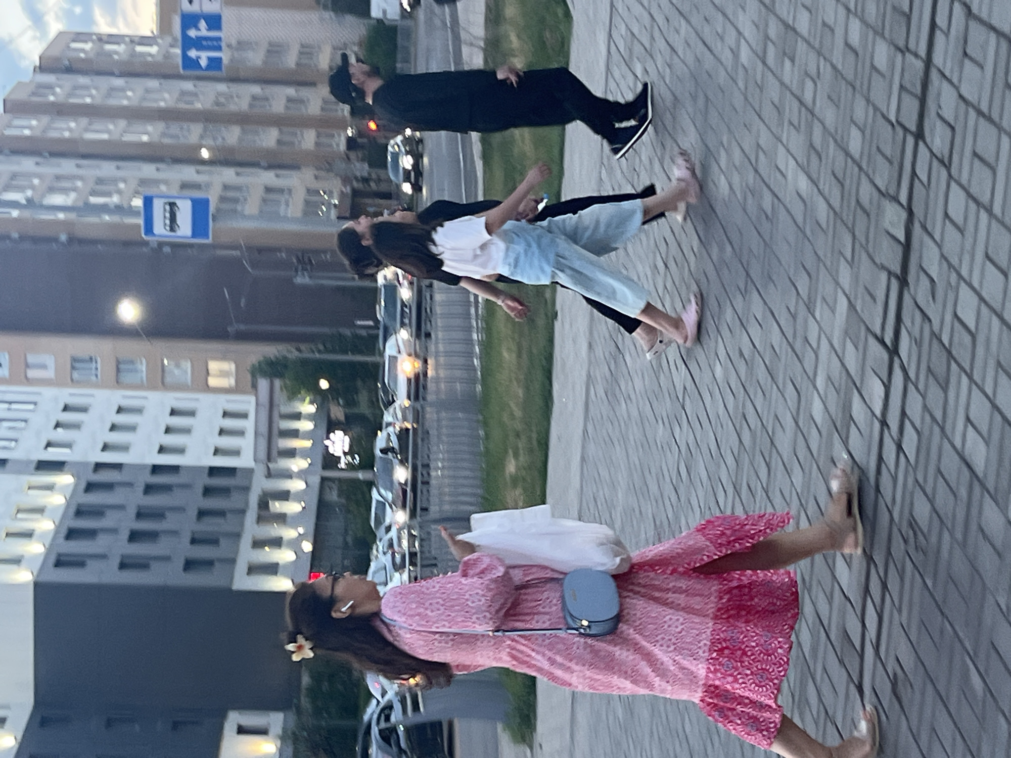 The image depicts a street scene in Astana, Kazakhstan, likely in the evening given the lighting conditions. A woman in a long, pink patterned dress and sandals walks from left to right across a paved sidewalk. She is carrying a white plastic bag in her right hand and a light blue crossbody bag over her left shoulder. She wears white earbuds. Behind her, two other individuals are walking in the same direction. The person in the middle, wearing a dark top and light blue culottes, is likely a younger female, followed by a person in a black outfit and a baseball cap.
In the background, modern apartment buildings with numerous windows and balconies are visible. A bus stop sign with a pictogram of a bus and a blue background is mounted on a pole. Traffic lights are present, and several cars are driving on the street, their headlights illuminated. The overall environment suggests a busy urban area in Astana, characterized by contemporary architecture and infrastructure.