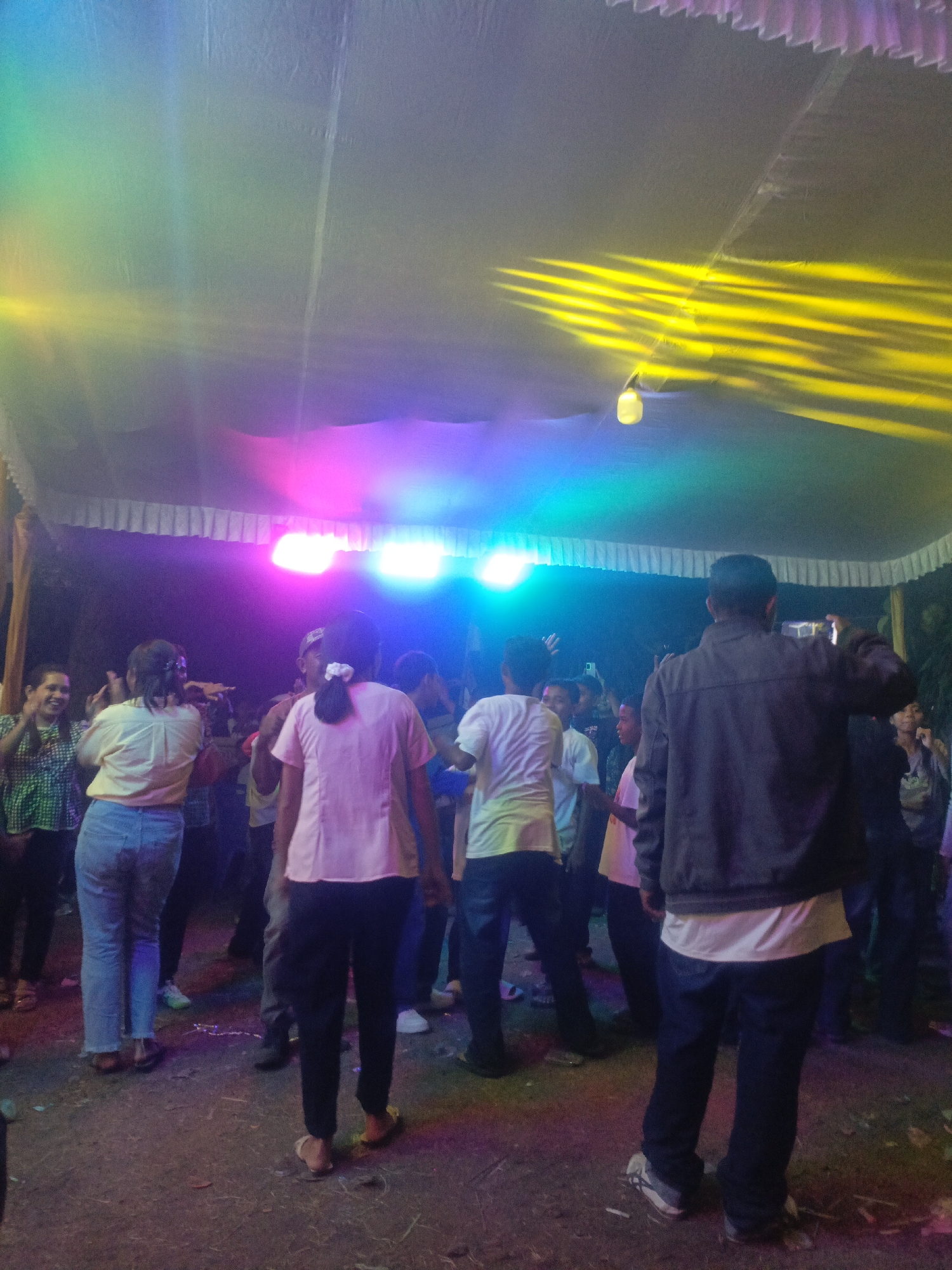 The image depicts a dimly lit outdoor gathering under a tent, possibly a party or celebration, with vibrant colored lights illuminating the scene. The location is specified as Solor Barat, Indonesia. Several individuals are visible, mostly from the waist down or with their backs to the camera, suggesting they are engaged in an activity such as dancing or socializing. The ground appears to be dirt, with some debris scattered around. The tent's fabric is illuminated by the colorful lights, creating a festive atmosphere. The overall impression is one of a lively community event taking place after dark in a tropical environment.