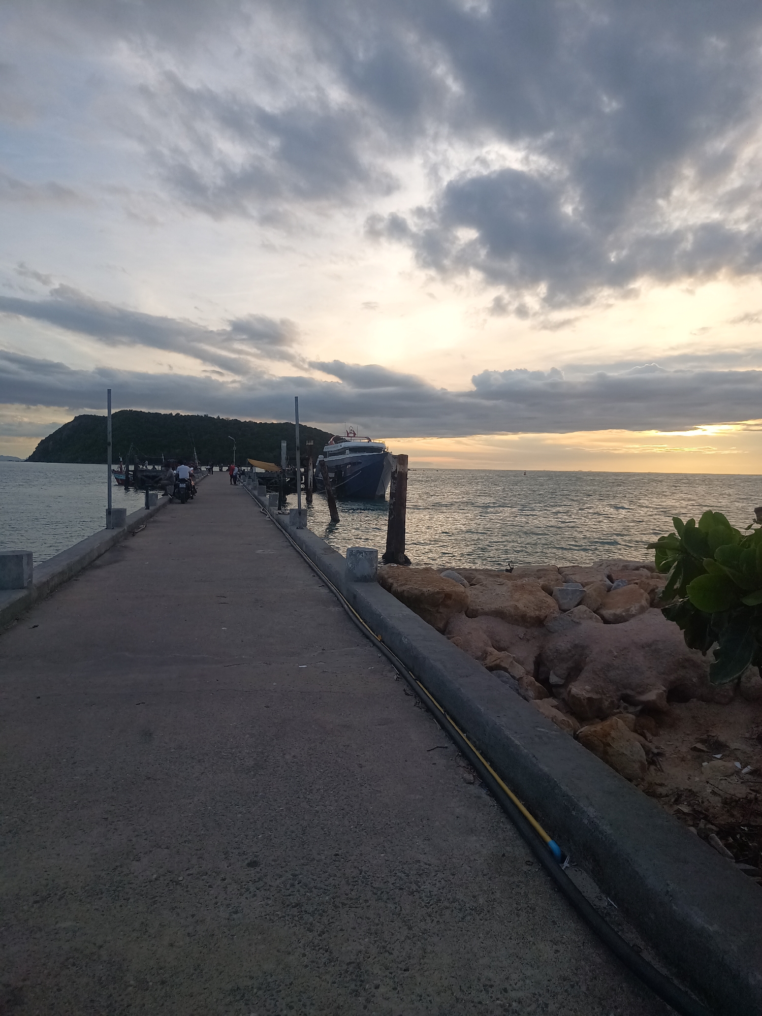 The image depicts a concrete pier stretching into the sea in ตำบลเกาะพะงัน, Thailand. A boat is docked on the right side of the pier, secured to wooden posts. Several people are walking on the pier in the distance. An island is visible in the background, and the sky is filled with clouds. To the right of the pier, large rocks are present with some greenery. Along the pier's edge, there's a concrete barrier with black and yellow cables running along it. The time appears to be late afternoon or early evening.