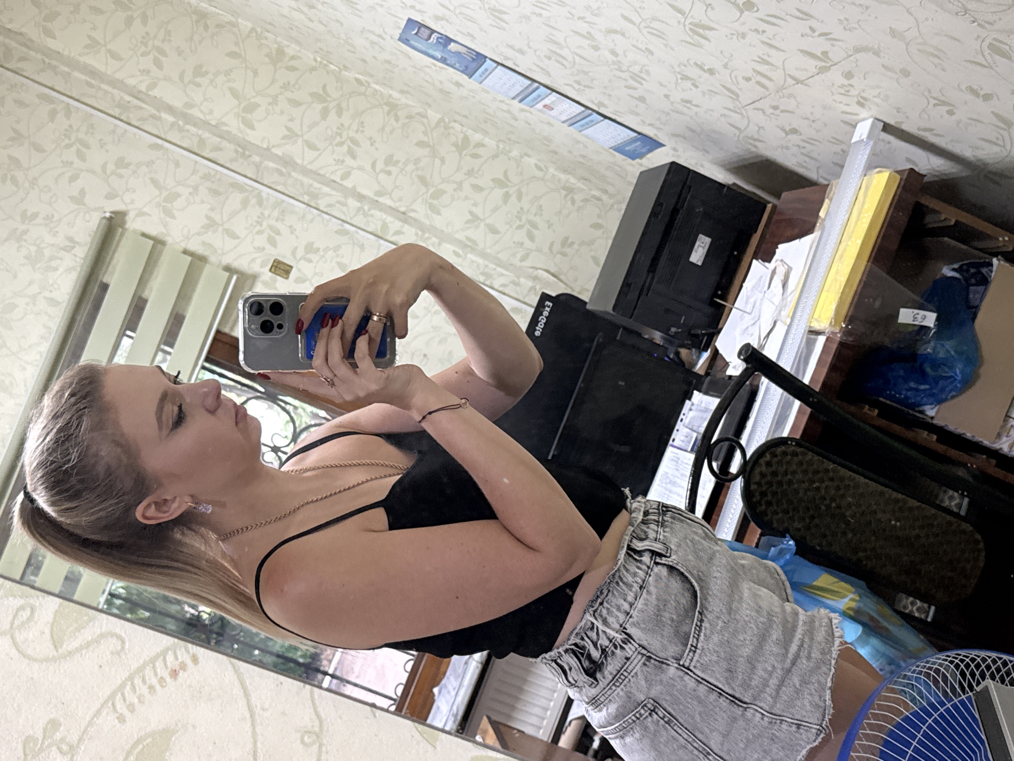 A young woman with blonde hair in a high ponytail takes a mirror selfie indoors. She is wearing a black spaghetti strap top and light-wash denim shorts. She has on a necklace, earrings, and rings, and her fingernails are painted red. She holds a smartphone in her right hand, angled to capture her reflection. The background shows a room with patterned wallpaper, a desk with a printer and papers, a fan, and a calendar on the wall.