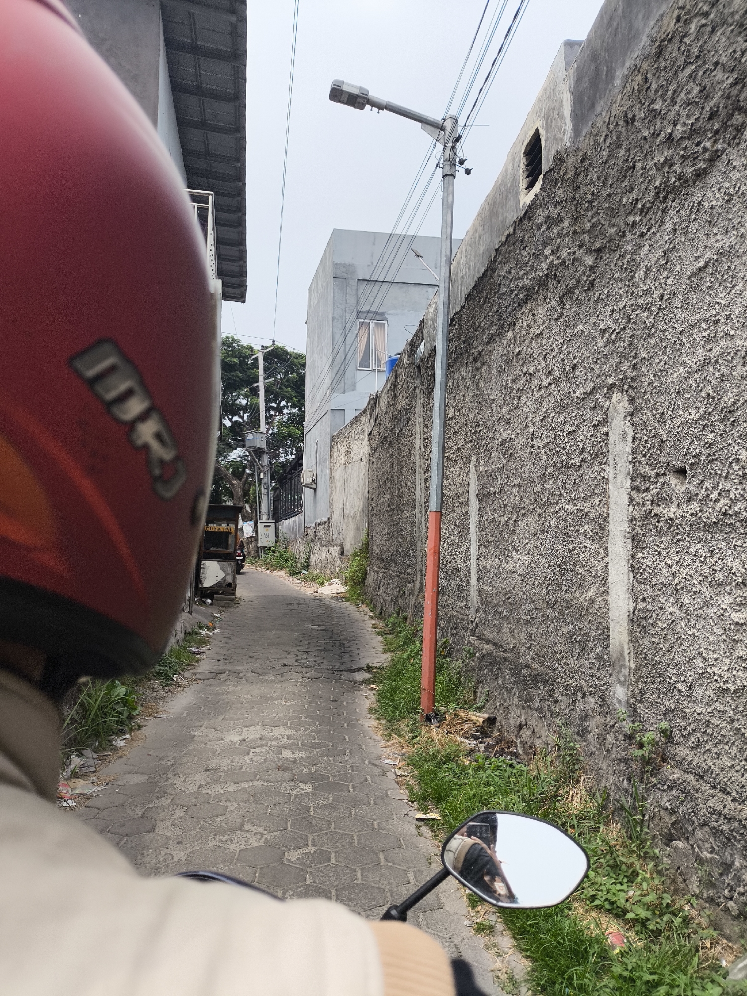 A first-person perspective shot from a motorcycle shows a narrow alleyway in Cilegon, Indonesia, with a textured concrete wall on the right and buildings on the left. A red motorcycle helmet is visible in the foreground, obscuring the rider's head. The alley is paved with hexagonal cobblestones and lined with sparse vegetation and debris. A lamppost stands in the middle of the alley, and a small food cart is parked on the left side. In the distance, another motorcycle is seen riding away. The sky is overcast. The wall on the right has a rough, stucco-like finish with some vertical cracks. A small, rectangular opening is visible near the top of the wall. The building on the left appears to be two stories high with a tiled roof. A side mirror of the motorcycle reflects a portion of the rider and the environment.
