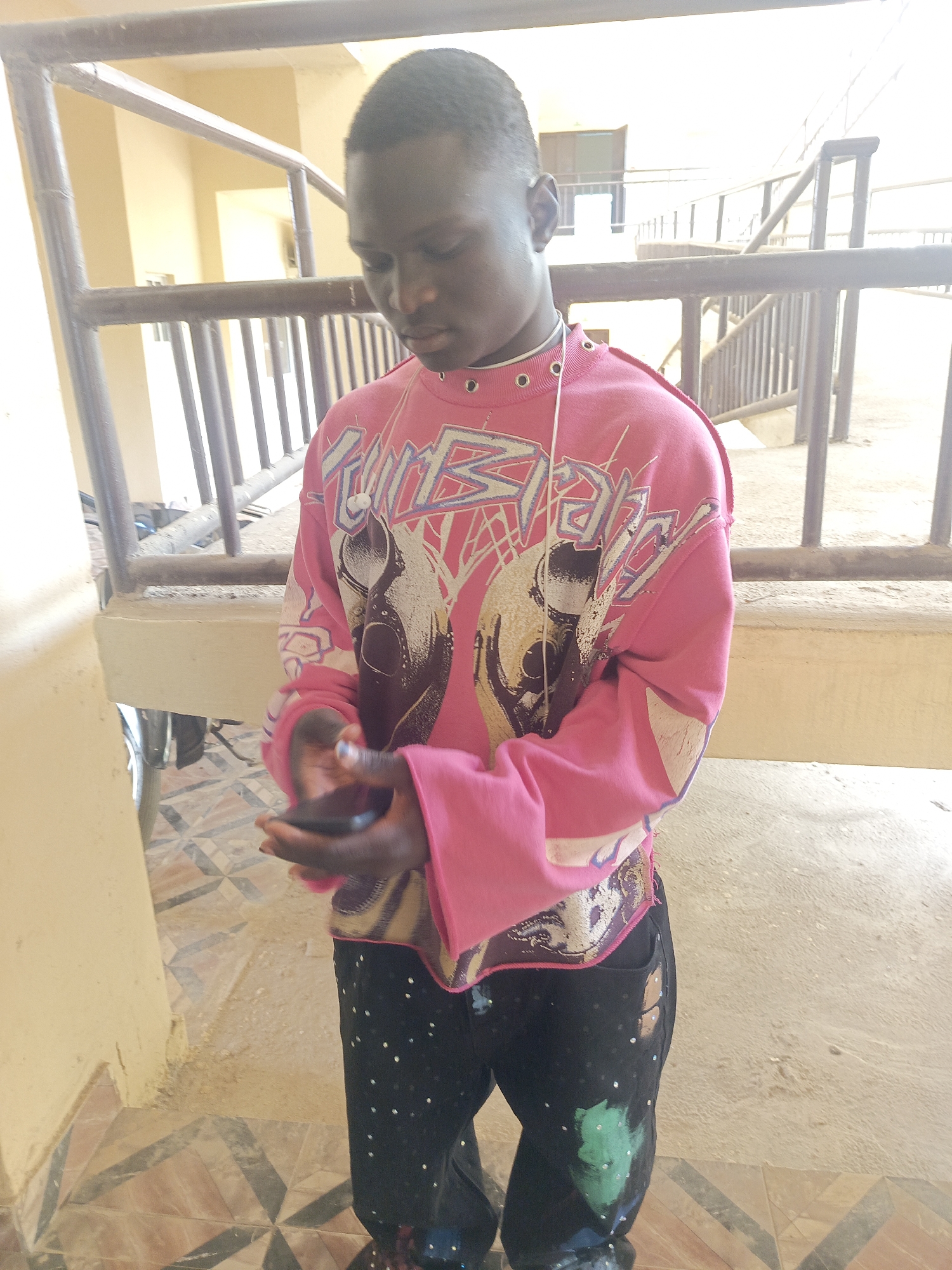 A young Black man stands on a tiled floor in Akwanga, Nigeria. He wears a pink sweatshirt with a graphic design and black pants with paint splatter. He is holding a dark-colored smartphone in his hands. The background features a railing and an indoor building structure with yellow walls. The lighting is bright and seems to be natural daylight.