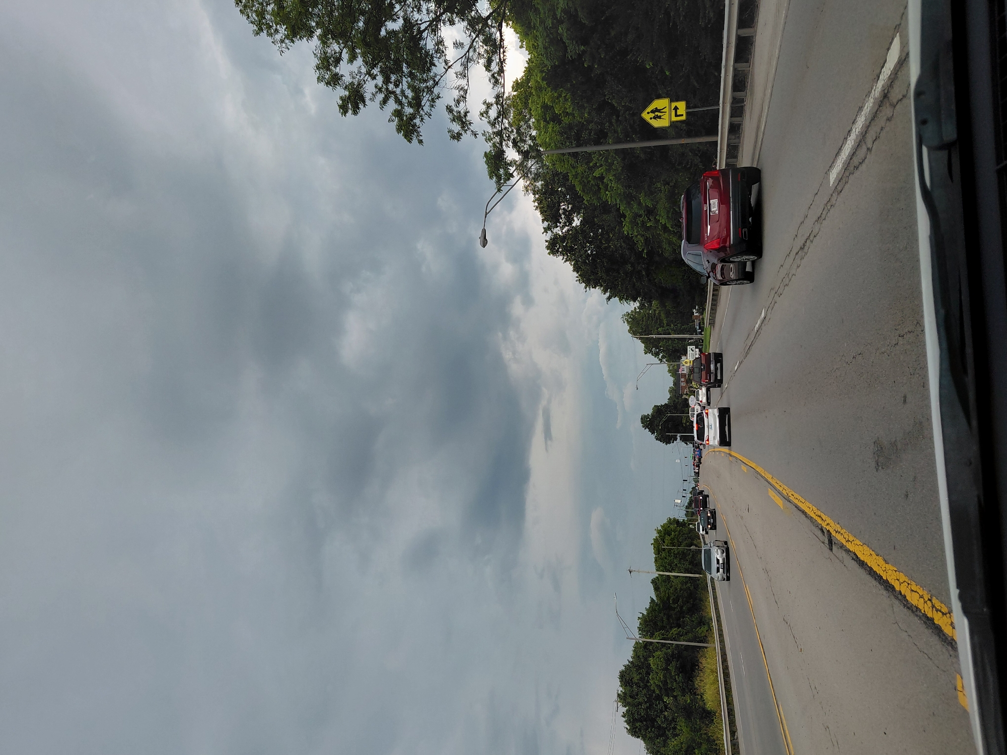 This is a vertical, eye-level, full shot of a highway in Lexington, United States of America, under a cloudy sky. The camera is positioned inside a vehicle, looking forward. The highway has two lanes in each direction, separated by a double yellow line. Traffic is moderate, with several cars visible. On the right side of the road, there is a guardrail and a sign depicting children crossing the street with a right-turn arrow. Trees line the right side of the highway, with more trees and buildings visible in the distance. The sky is overcast with dark, heavy clouds, suggesting impending rain.