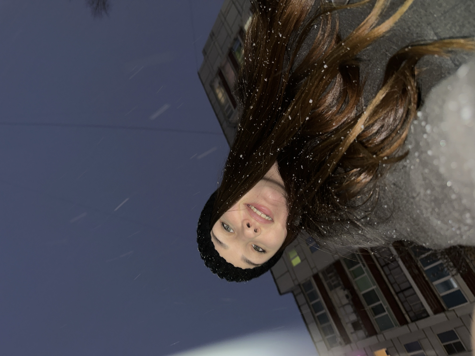 A young woman is pictured outdoors, likely in the city of Chaikovsky, Russia, given the architectural style of the building behind her. It appears to be snowing lightly, as snowflakes are visible on her hair and clothes, and in the air. The time of day is either very early morning or late evening, as the sky is a dark blue/grey color. The woman is wearing a dark knitted beanie and has long, brown hair. She's looking at the camera with a slight smile. The building in the background is multi-storied with windows illuminated from within. The photo is taken from a low angle, looking up.