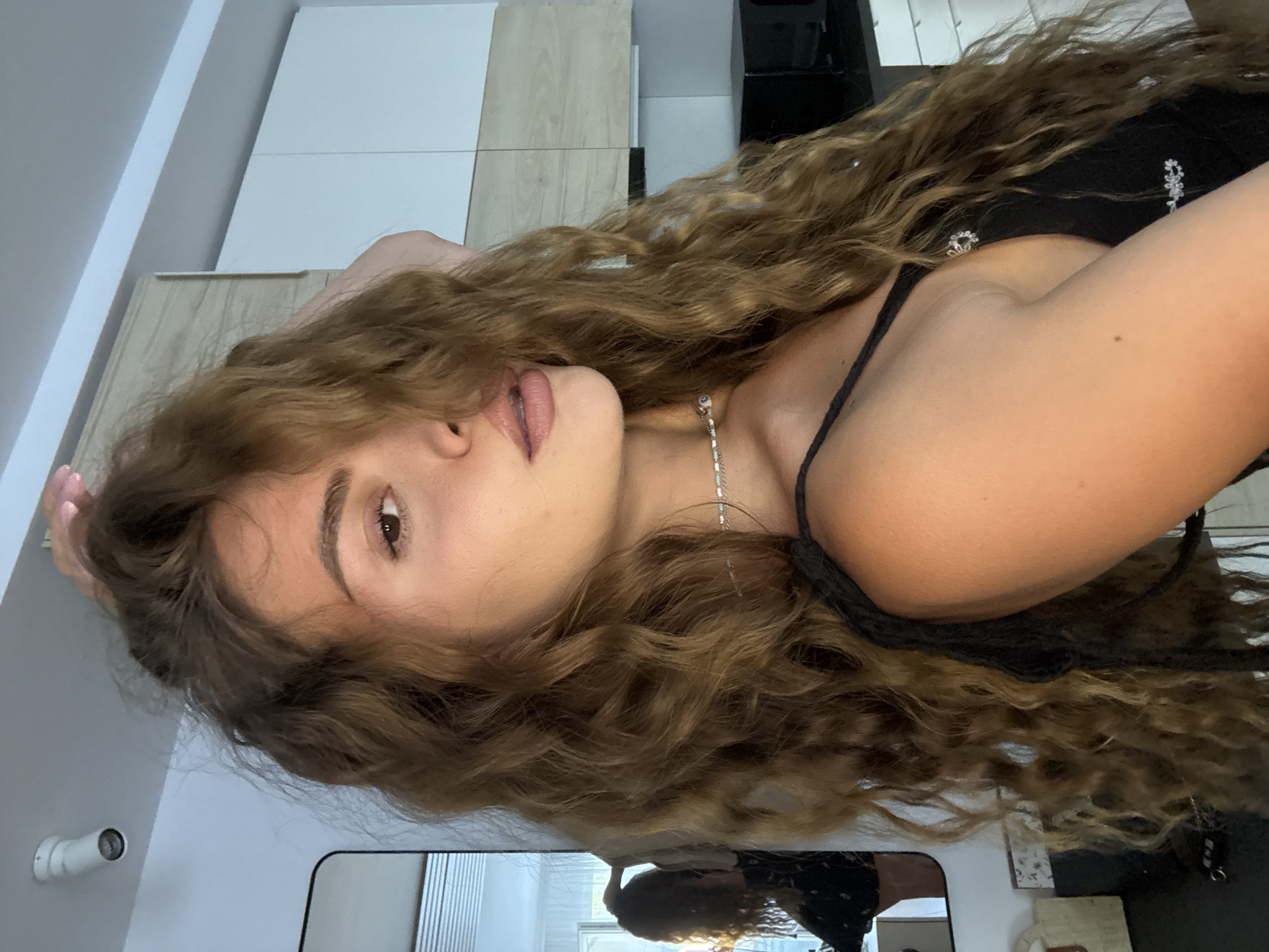 A tanned, young woman with long, curly brown hair is the central figure in this close-up shot. She's wearing a black spaghetti strap top and a silver necklace. Her makeup is subtle, featuring lip gloss and defined eyebrows. She is positioned indoors. The background includes white and light wood-colored cabinets. A black object, possibly a refrigerator or storage unit, is visible on the upper right. A mirror is located in the lower portion of the image. The location of this photograph is the city of Unknown, Unknown.