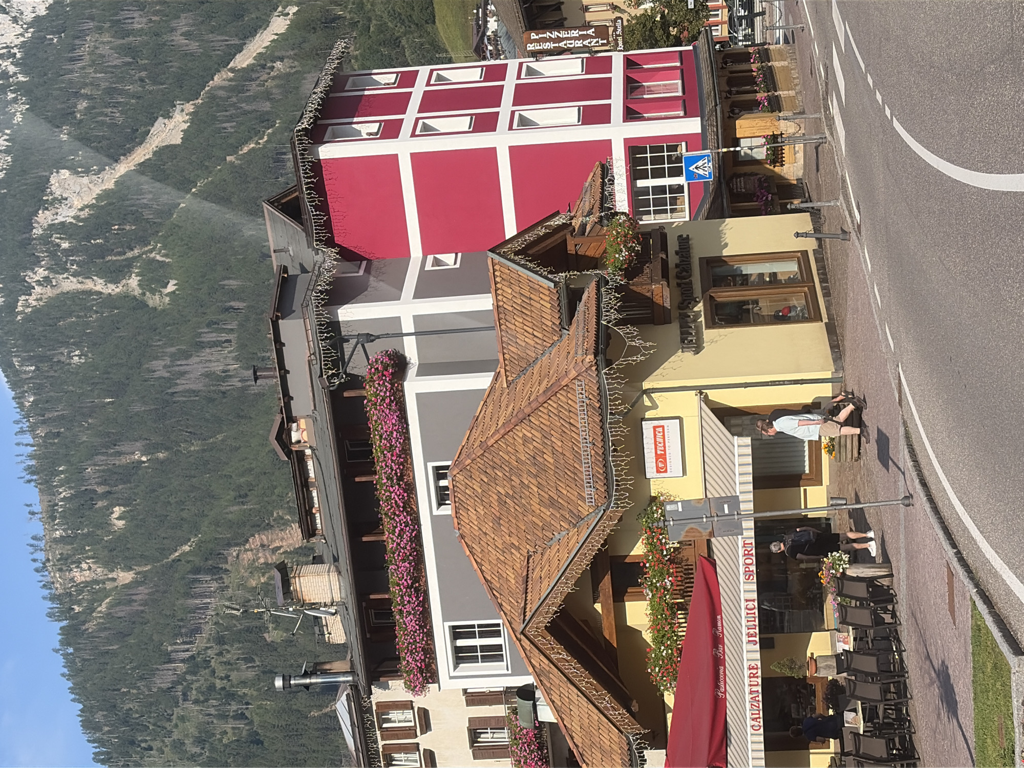 In Moena, Italy, a street scene unfolds with buildings and a mountain backdrop. A bright red, multi-story building stands prominently, contrasting with the adjacent grey structure. In front, a yellow building features shops with awnings. One awning displays "PASTICCERIA BAR RAMON," and another has "CALZATURE JELLICI SPORT" printed on it. Several tables and chairs are set up outside a cafe, with a few individuals walking on the sidewalk. The street curves in the foreground, with a pedestrian crossing marked. The background is dominated by a steep, tree-covered mountain slope, indicating the alpine setting of Moena.