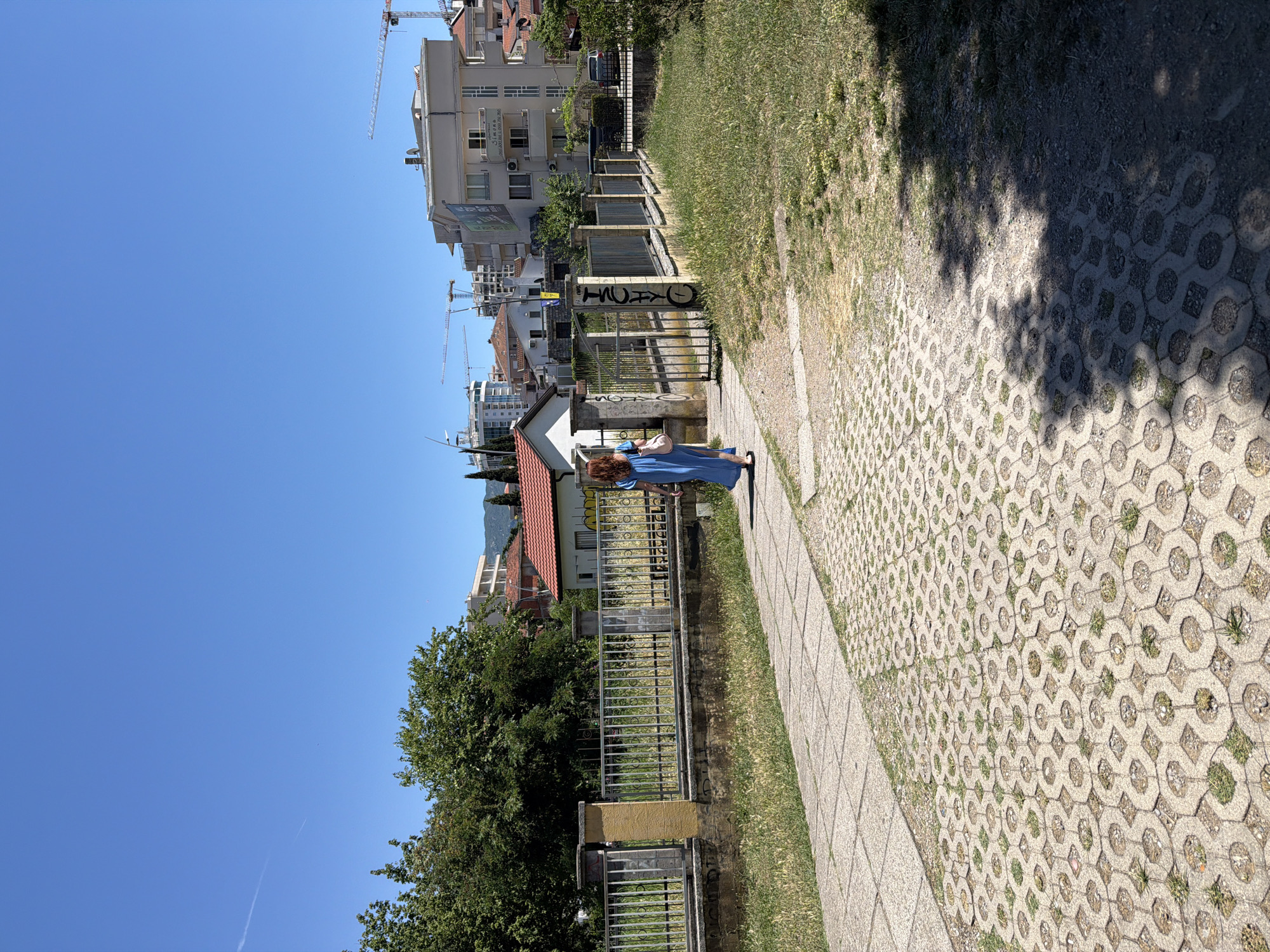 A woman wearing a blue dress walks along a paved pathway in Budva, Montenegro, on a sunny day. The pathway is bordered by a grassy area and a metal fence. Behind the fence, a small building with a red roof is visible, as well as several apartment buildings and construction cranes in the distance. The sky is clear and blue.