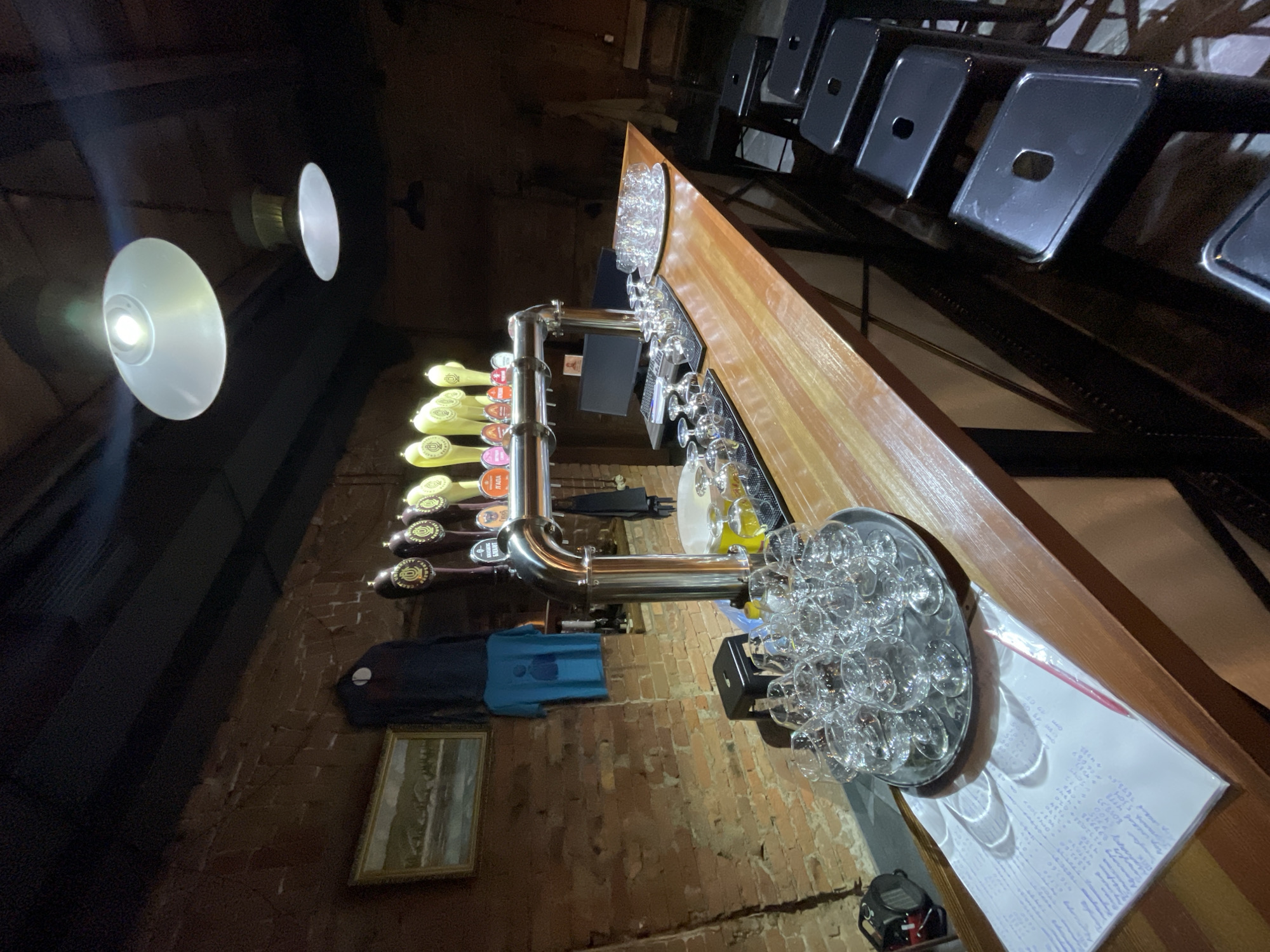 The image shows the interior of a bar or pub, likely located in Krasnoyarsk, Russia, featuring a beer tap system mounted on a brick wall. The tap system has multiple beer taps with different handles. A wooden bar top extends horizontally, laden with glassware and a menu or order pad with handwriting. Above the bar, seating is arranged, with metal backrests. Two prominent light fixtures are visible, casting light on the scene. The environment appears dimly lit, with exposed brick and industrial-style elements. A framed landscape painting hangs on the wall near the beer taps. There are no people visible.