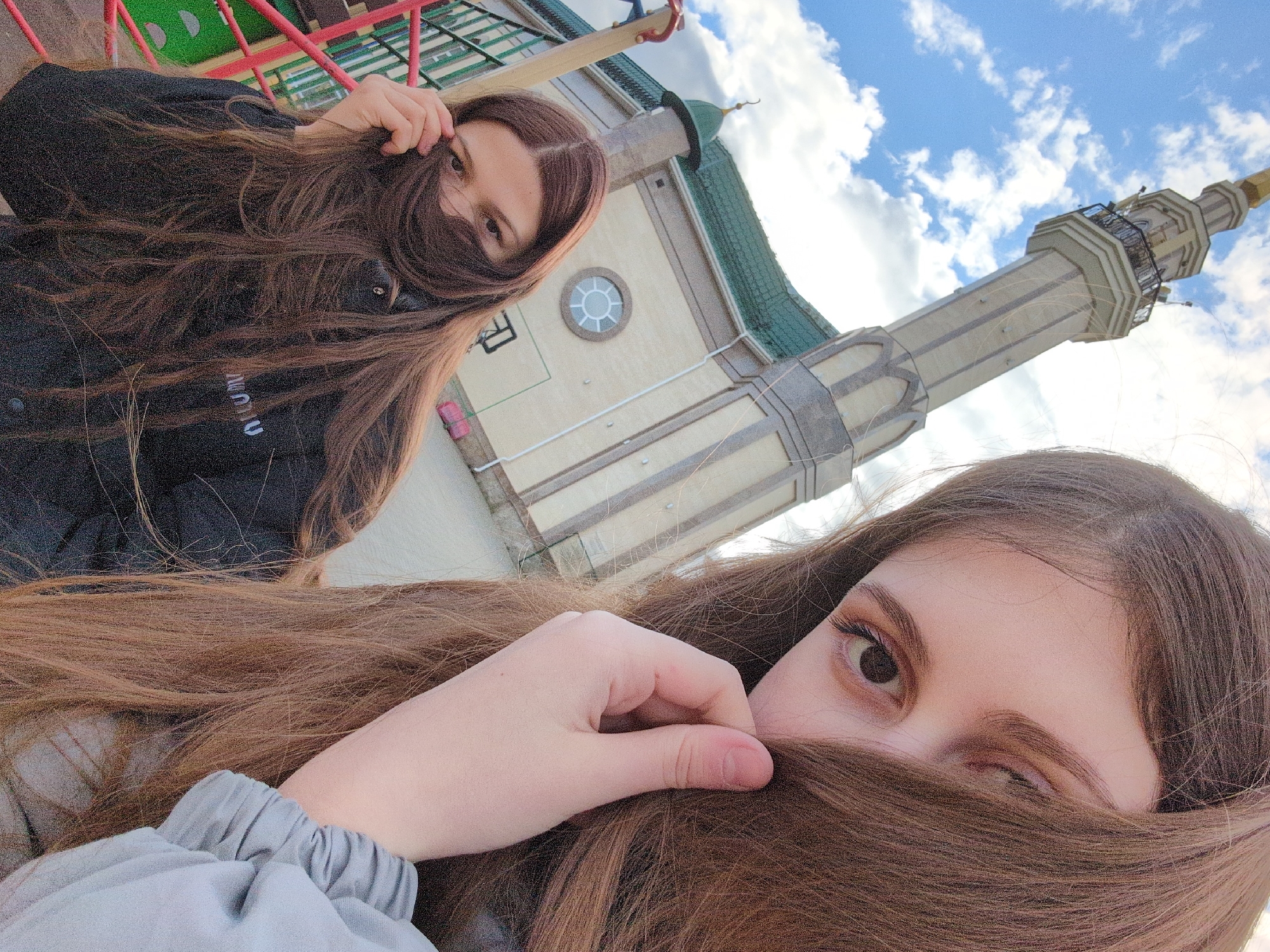 Two women are pictured in this upward-angled shot against a backdrop of a light beige building with a green-tiled roof and a tall, ornate minaret reaching towards a cloudy blue sky. The building's architectural details include a round window with radial glazing and horizontal banding. The minaret features multiple tiers, each with a balcony, and culminates in a gold-colored spire.
The woman in the foreground partially obscures her face with long, brown hair, with one eye visible and a hand near her face. She wears a light gray garment. The other woman, positioned further back, also has brown hair partially covering her face. She is dressed in a black jacket. A colorful playground structure is visible in the upper left corner of the frame.
The scene is located in Izhevsk, Russia, as evidenced by the distinctive architecture of the minaret, which is associated with the Izhevsk Mosque.