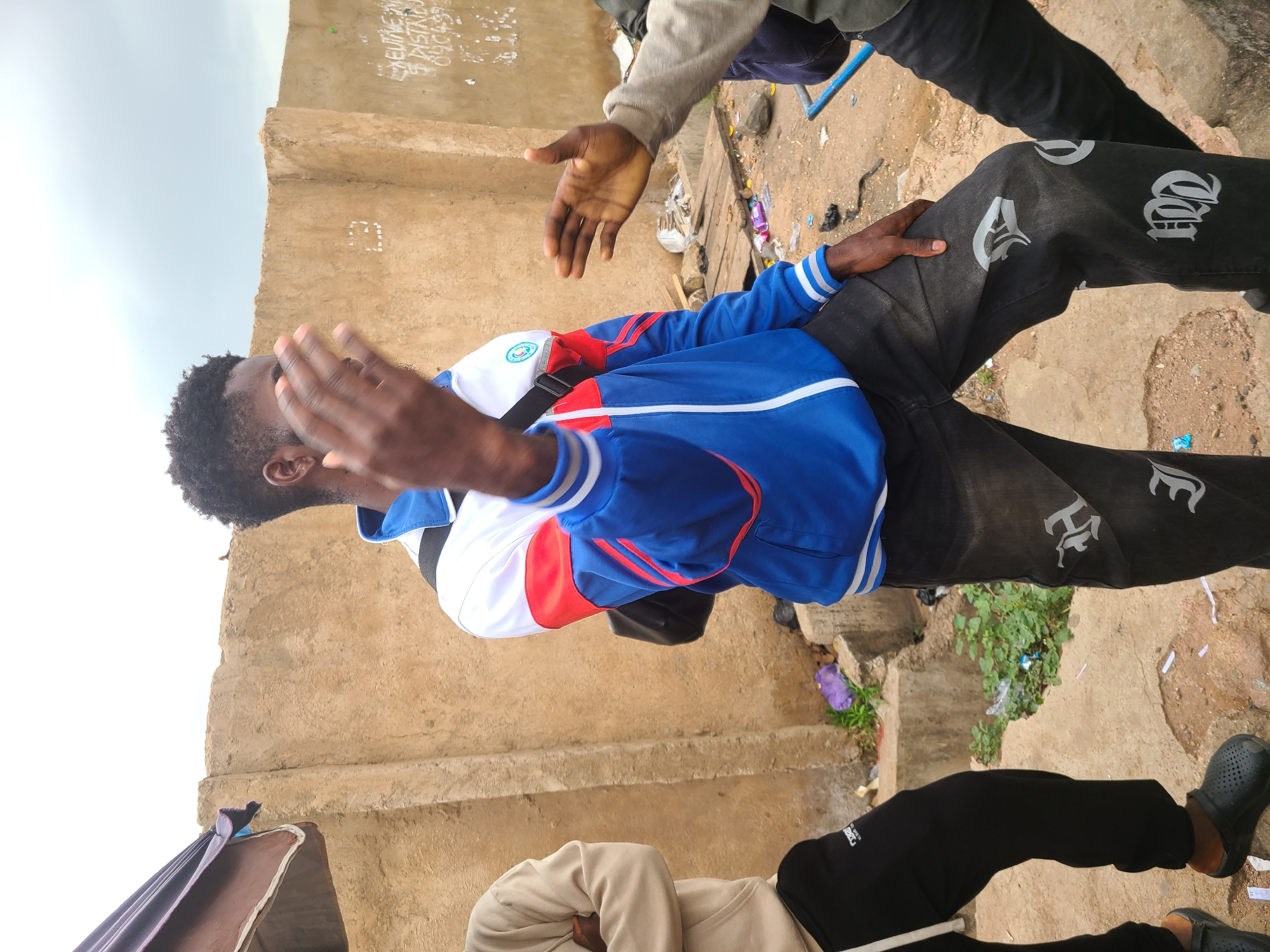 In Gura Top, Nigeria, two individuals are captured in an outdoor scene. One person, predominantly in focus, wears a blue and white jacket and black jeans with light-colored designs. This person's hand is raised. Another person is positioned partially in the frame with a gray sweatshirt and black pants. A tall, tan-colored building appears in the background, marked with what seems to be writing or graffiti. The ground appears to be a concrete surface with litter and vegetation.