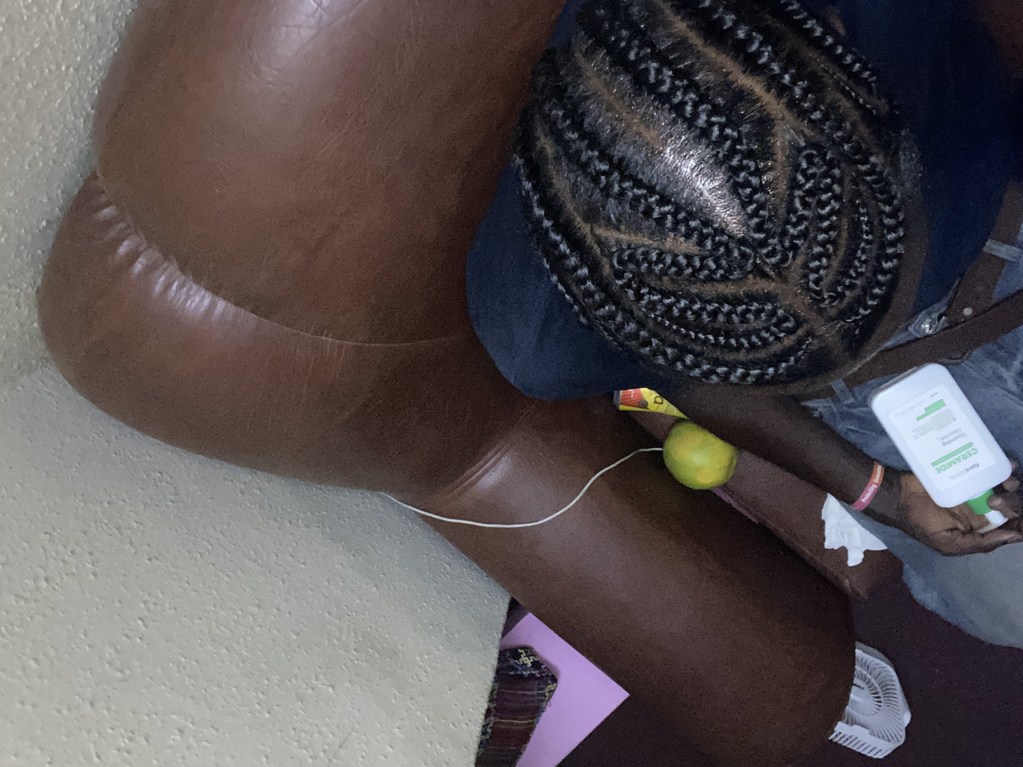 An overhead image depicts an individual seated in an indoor setting. The person, with dark skin, wears blue jeans and has black hair styled in intricate cornrows revealing the scalp. A pink wristband is visible on their left arm. In their right hand, they hold a white bottle with a green cap, labeled "CERAMIDE Foaming Cleanser." A brown strap, likely from a bag, crosses their shoulder area. To the left, a large, dark brown, upholstered furniture piece is visible, upon which rests a single green-yellow citrus fruit and a thin white cable. A white textured wall is in the upper left background. On the floor in the lower left, a small white USB fan is present, alongside a pink sheet of paper and a patterned fabric item.